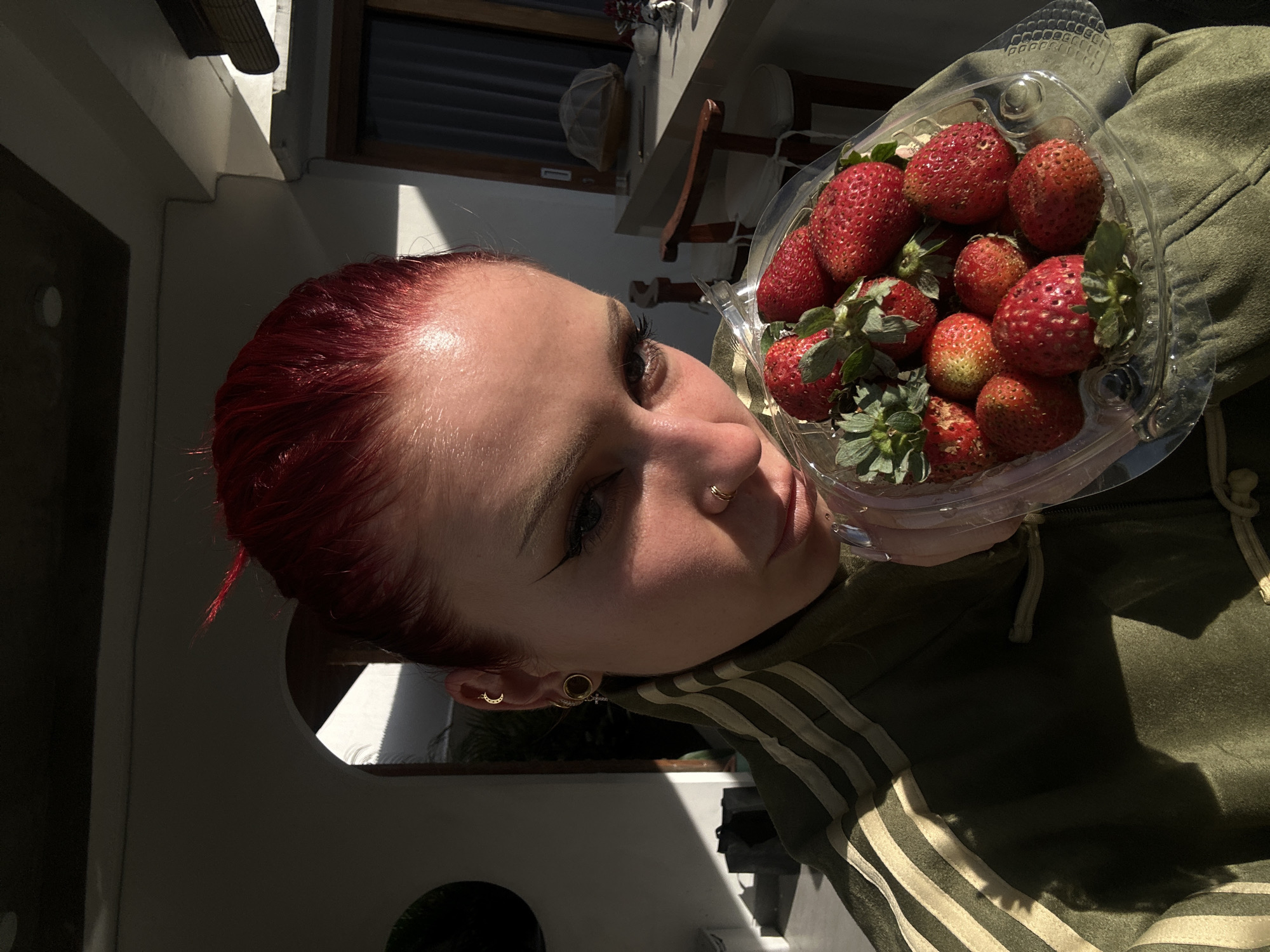 A fair-skinned individual with dyed red hair is holding a clear plastic container of strawberries. The person has a nose ring and multiple ear piercings. They are wearing a green and white striped hoodie. The background includes part of a building exterior with a window and an archway. The scene is outdoors in North Kuta, Indonesia, during the daytime, as evidenced by the natural lighting.