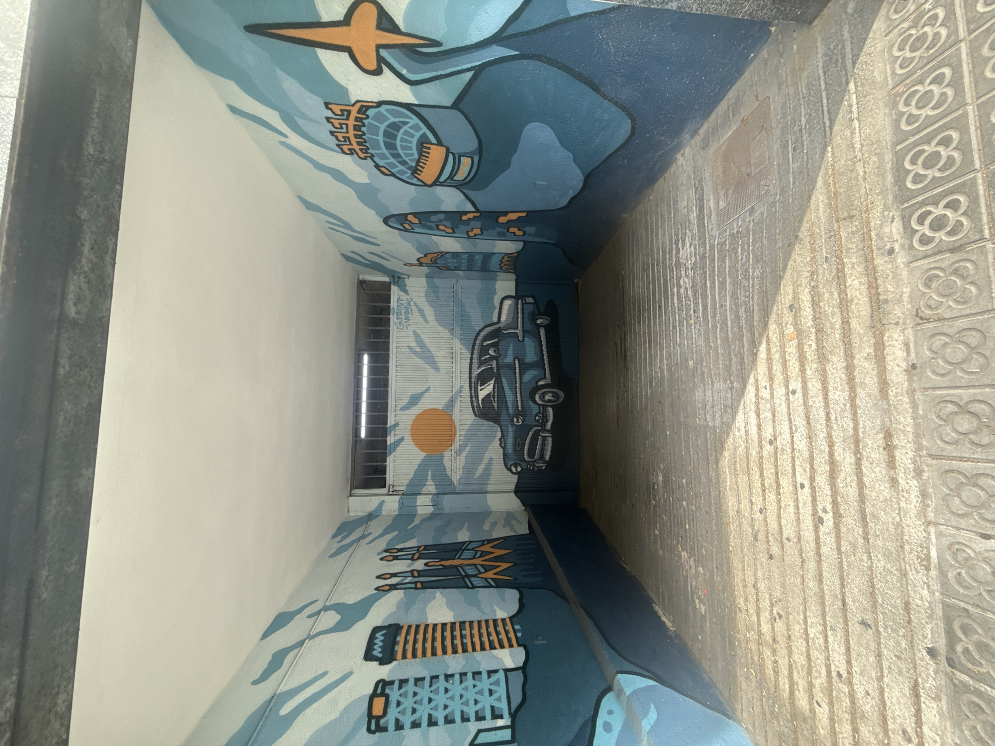 The image shows a narrow, indoor tunnel with a mural painted on the walls. The mural depicts a stylized cityscape with buildings and a vintage car. The style of the buildings appears to be inspired by iconic Barcelona architecture, such as Sagrada Familia, and modern skyscrapers. The color palette consists mainly of blues, oranges, and grays. The tunnel floor is made of concrete with visible lines, suggesting a walkway or ramp, and decorative tiles are visible at the bottom of the image. The ceiling of the tunnel is painted white. The overall impression is that of an urban underpass or passage decorated with art. The mural on the wall includes the signature "GAINT PAINT - WORK."