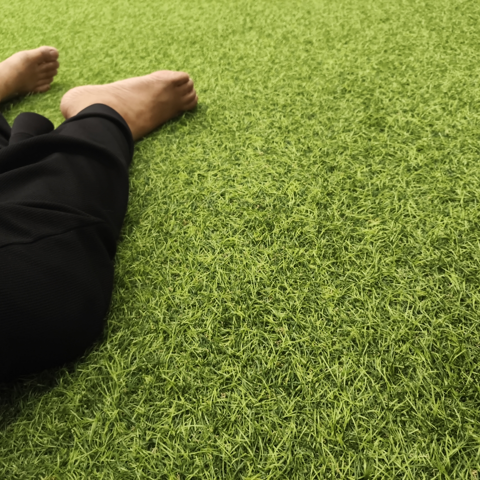The image shows a close-up view of two bare feet lying on bright green artificial turf. The feet are positioned in the lower left quadrant of the frame, with the toes of one foot visible and the sole of the other. Black pants are visible on the left side, partially covering the legs. The artificial turf fills the majority of the frame, creating a textured, green background. There is no information in the image to indicate a specific location in South Jakarta, Indonesia.