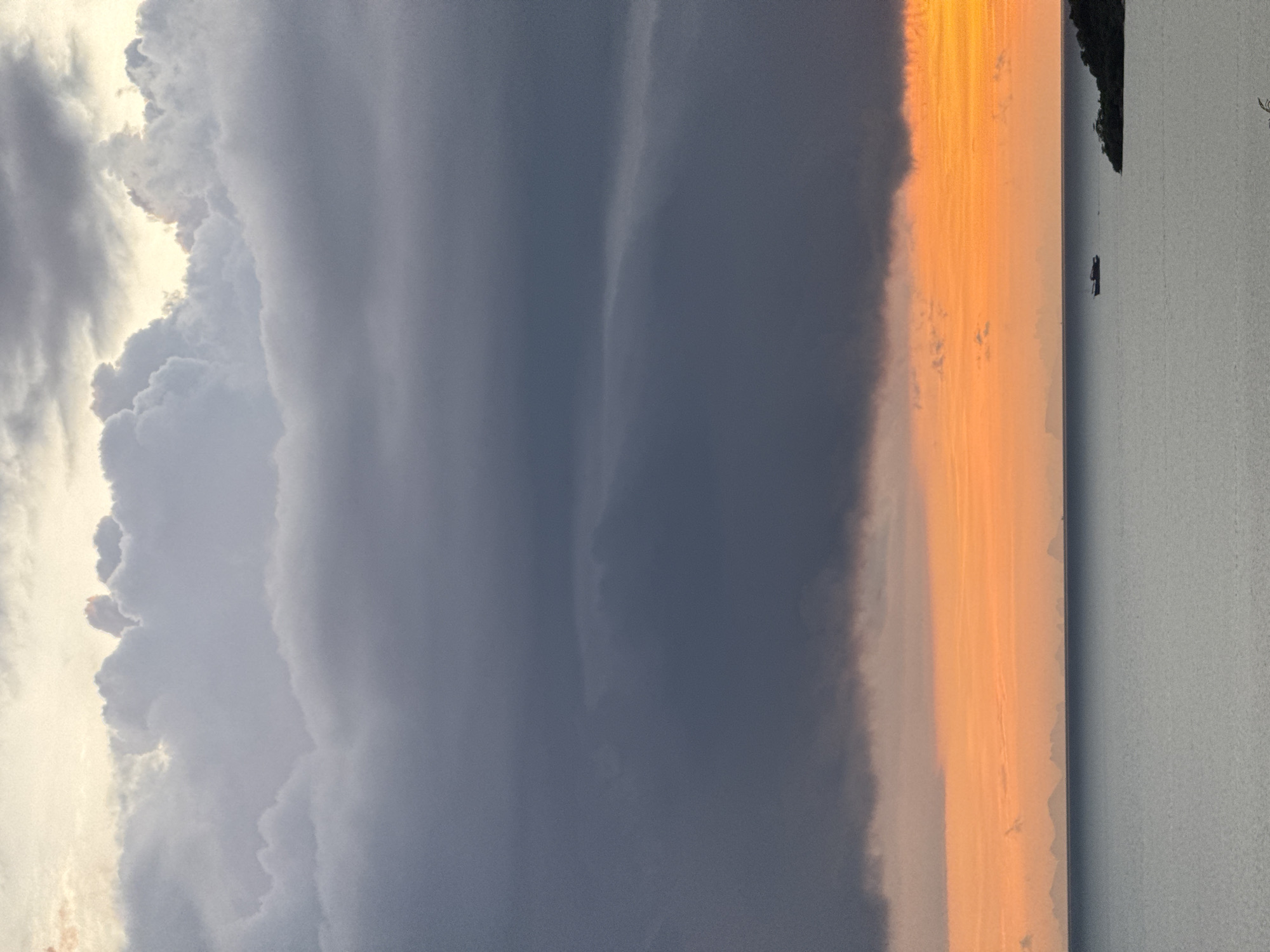 A vertical shot captures the sky and a body of water at dusk. The sky is dominated by dramatic clouds, with lighter, fluffy clouds at the top transitioning to darker, more ominous clouds towards the horizon. Below the dark clouds, a band of vibrant orange and yellow illuminates the sky, indicative of sunset. The water is calm and reflects the colors of the sky. In the distance, a faint outline of land or islands can be seen along the horizon. A small boat is visible in the middle of the water, and on the right side of the frame, a dark green, tree-covered landmass juts out into the water. The overall impression is a serene yet powerful natural scene.