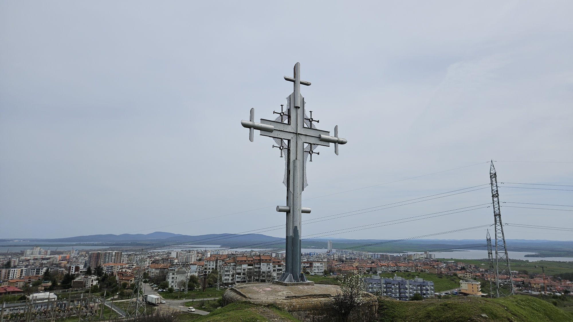 The image shows a large metal cross atop a grassy hill overlooking the city of Burgas, Bulgaria. In the background, the cityscape stretches towards a body of water with hills beyond. Power lines and a utility tower are visible to the right of the cross. The sky is overcast. The cross is mounted on a concrete base.
