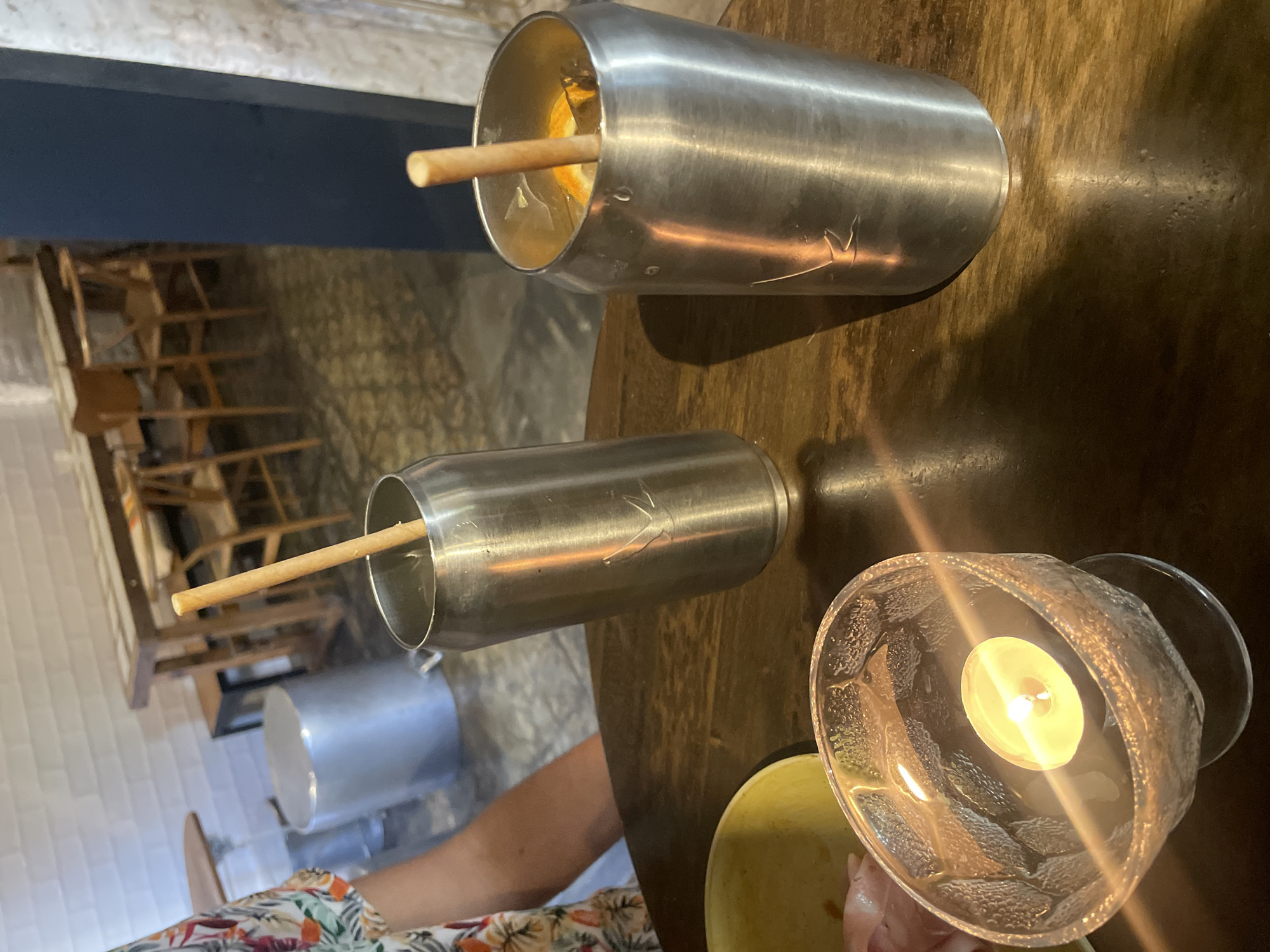 The image shows a close-up, angled shot of a wooden table setting, likely in a restaurant or bar. The angle is slightly askew, with the top-right portion being higher than the bottom-left.
Two cylindrical metal containers, possibly cocktail glasses or cups, lie horizontally on the table. Each has a straw sticking out. The one on the right appears to contain liquid and perhaps fruit. To the bottom right of the frame is a decorative glass candle holder with a lit candle inside.
A section of a person’s arm and floral-patterned shirt are visible at the bottom of the frame.
The background includes a glimpse of seating arrangements and a wall, suggesting an indoor setting. Based on the style and location, it's plausible that this scene is in Barcelona, Spain, a city known for its vibrant culinary scene and unique presentation styles.