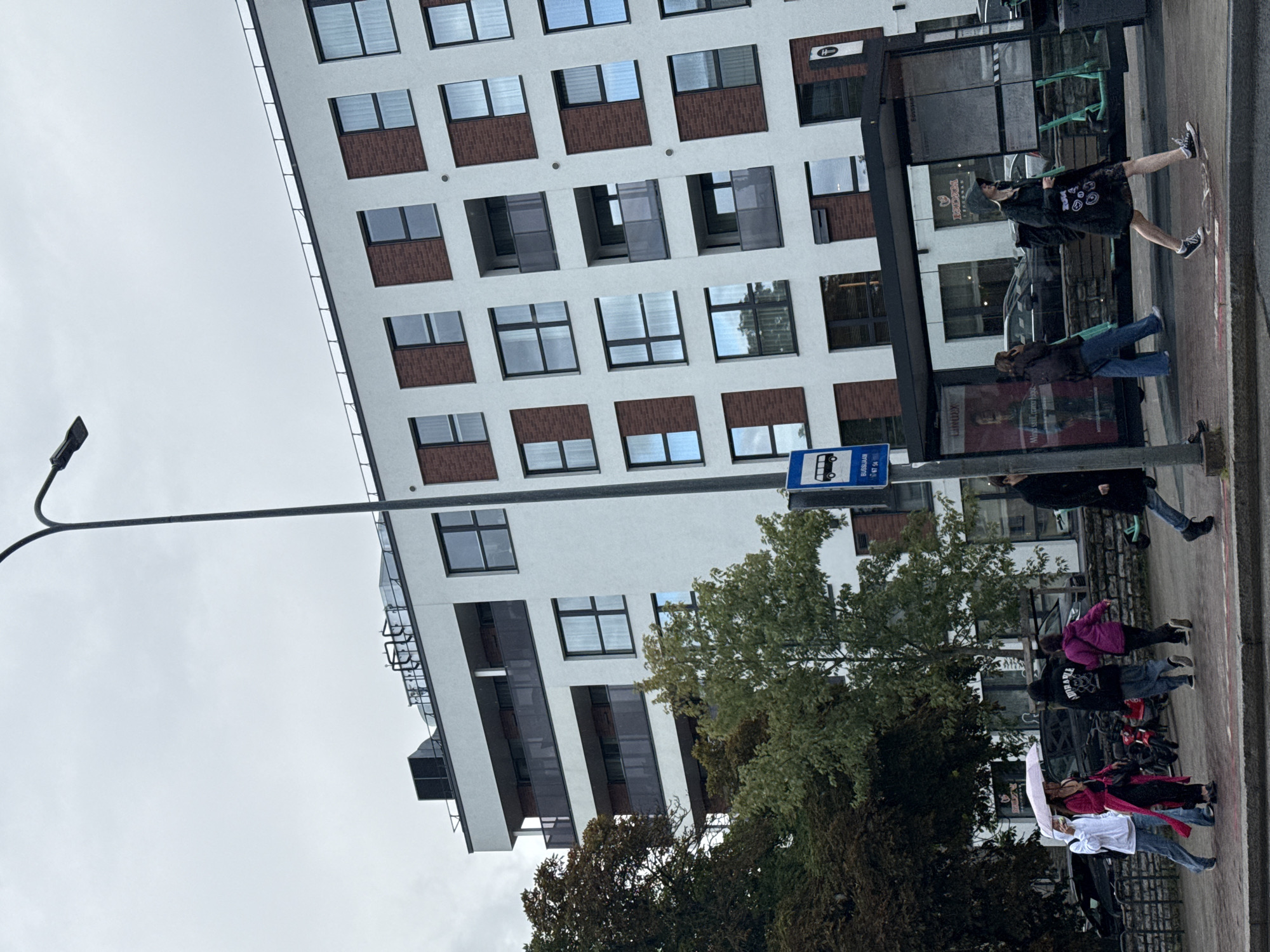 This is a medium-shot, eye-level photograph of a street scene in Tallinn, Estonia, under an overcast sky. In the foreground, a paved sidewalk runs horizontally across the frame. Several people are walking on the sidewalk. On the left, three individuals are walking together; one person on the far left is wearing a white hoodie and blue jeans, holding a pink umbrella over their head and walking with another person dressed in a bright pink long coat and black pants. Behind them, a third person in a black hoodie and blue jeans is also walking. Further to the right, another person in a dark purple jacket and black pants is walking. On the right side of the frame, a person in a black coat and dark pants is walking away from the camera.
Behind the sidewalk, a modern apartment building with a white facade and red brick accents dominates the background. The building has multiple floors with many windows and balconies. A street lamp with a dark pole stands tall on the left side of the frame, extending into the upper left corner. Adjacent to the building is a bus stop shelter with a black frame and transparent walls, displaying advertisements and schedules inside. Several electric scooters are parked outside the bus stop. Trees with green and brown foliage are visible on the left side of the frame, partially obscuring the lower floors of the apartment building. A bus stop sign with a blue background and a white bus icon is posted on a pole in front of the bus stop shelter. The overall impression is of an urban environment, possibly on a slightly damp or rainy day given the umbrella.