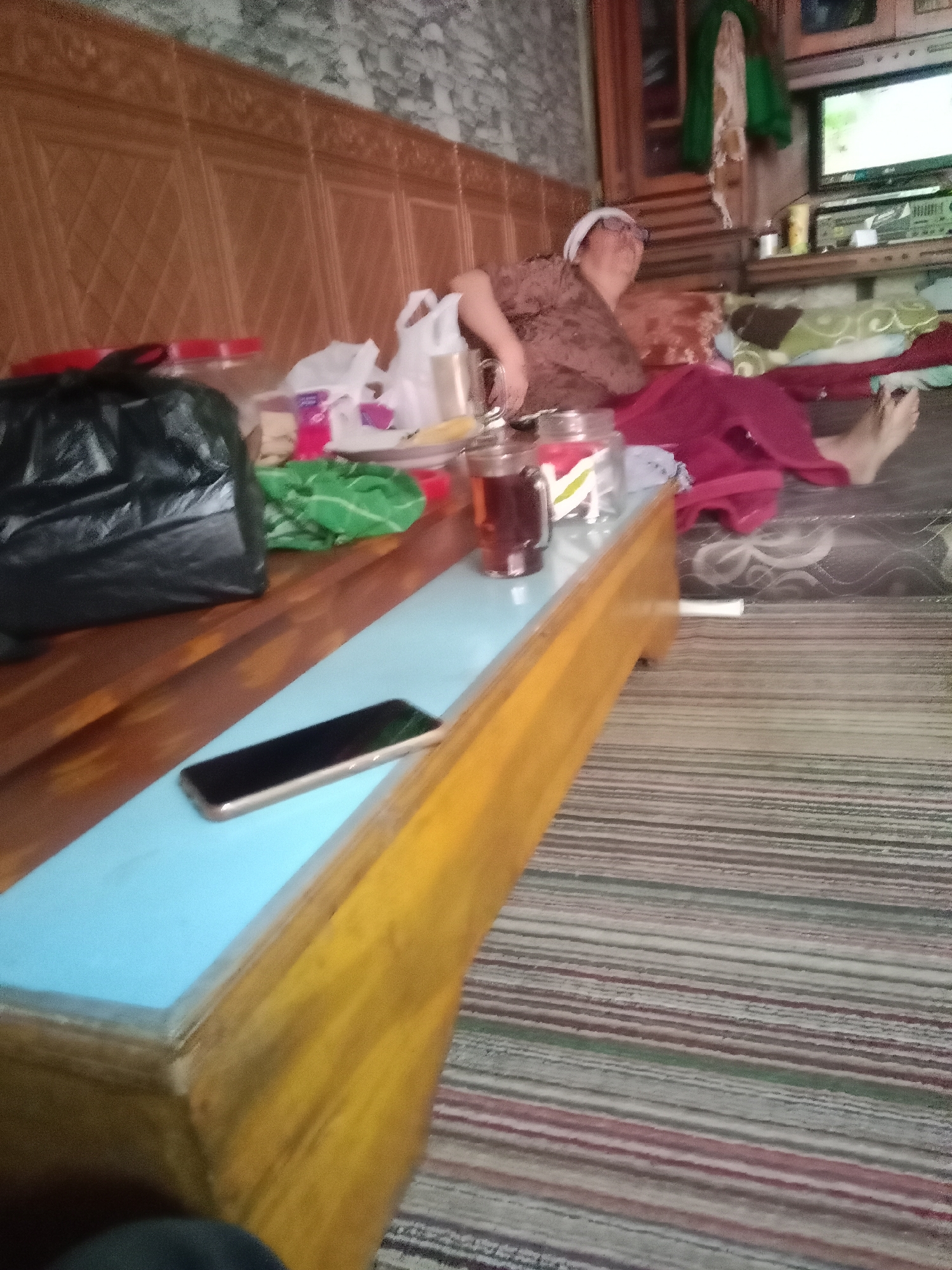 The image shows an indoor scene, likely a living room or bedroom, with a person lying on a mattress. The person, who appears to be an adult, is wearing glasses and a brown patterned shirt with a white head covering. They are reclining on a mattress with a striped blanket spread over them. To the left of the mattress, there is a wooden table with a light blue surface. On this table, a black plastic bag, a green cloth, and several containers, including one with a dark liquid that appears to be tea, are visible. A smartphone is also placed on the blue surface of the table. In the background, a television set is on a shelf, and there are wooden wall panels. The overall impression is that of a casual, domestic setting in Patakbanteng, Indonesia.