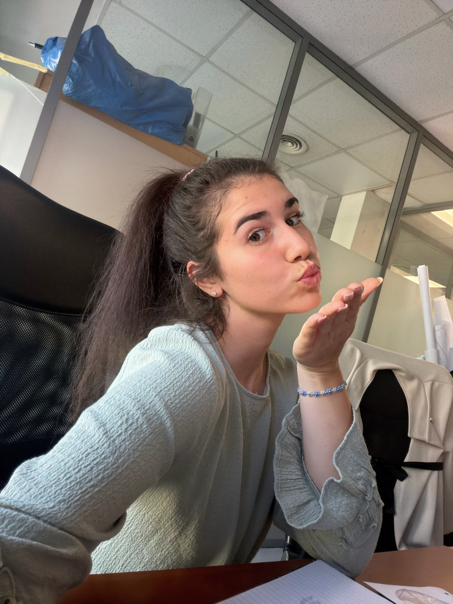 A young woman with dark hair pulled into a high ponytail, wearing a light blue textured shirt and a blue and white beaded bracelet, is seated in a black office chair. She is leaning slightly forward, holding her right hand up to her face with her palm extended outwards, as if blowing a kiss. Her lips are puckered, and her eyes are looking directly at the camera. She has a small stud earring in her visible ear.
The background suggests an office environment, with a ceiling of acoustic tiles and a glass partition with dark framing. Sunlight is streaming in from an unseen window, casting shadows and highlighting textures in the scene. On a shelf above and behind the woman, a blue tarp covers an object. A light-colored jacket or coat is draped over the back of another chair to the right of the woman. The desk surface in front of her is a dark wood color, with a notebook and papers visible.
The image does not contain any visual information that would allow for the identification of the city of Sofia, Bulgaria.