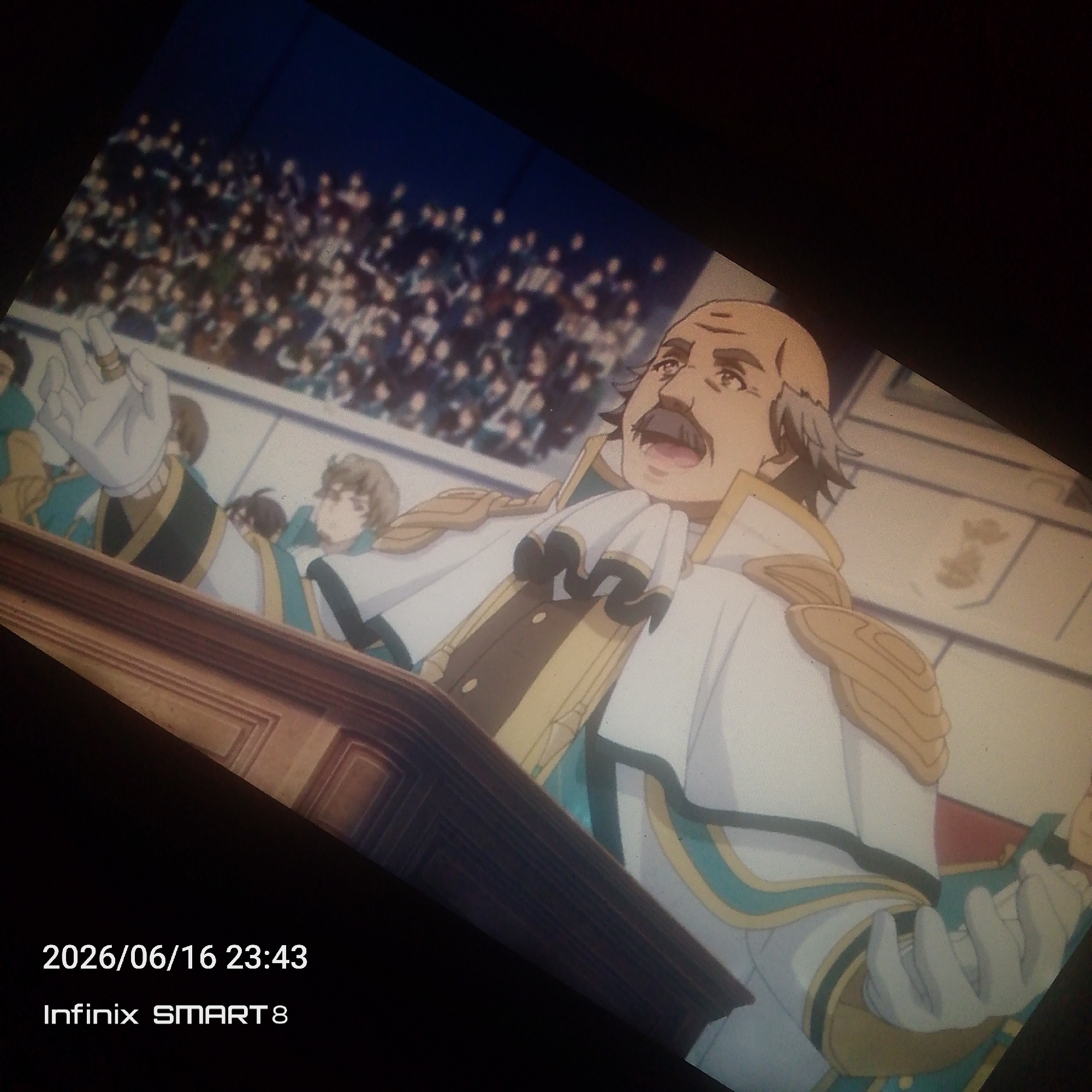The image shows an anime-style depiction of a man with a mustache and balding head, wearing a formal white and gold uniform with a large ruffled collar. He is standing behind a wooden podium, with his mouth open as if speaking. In the background, a crowd of people are visible in tiered seating, and to the left of the man, several younger figures in similar uniforms are also present. The scene appears to be set indoors, possibly in an auditorium or a formal hall. There is a timestamp in the bottom left corner indicating "2026/06/16 23:43" and "Infinix SMART 8". There is no information in the image that relates to the city of Jos, Nigeria.