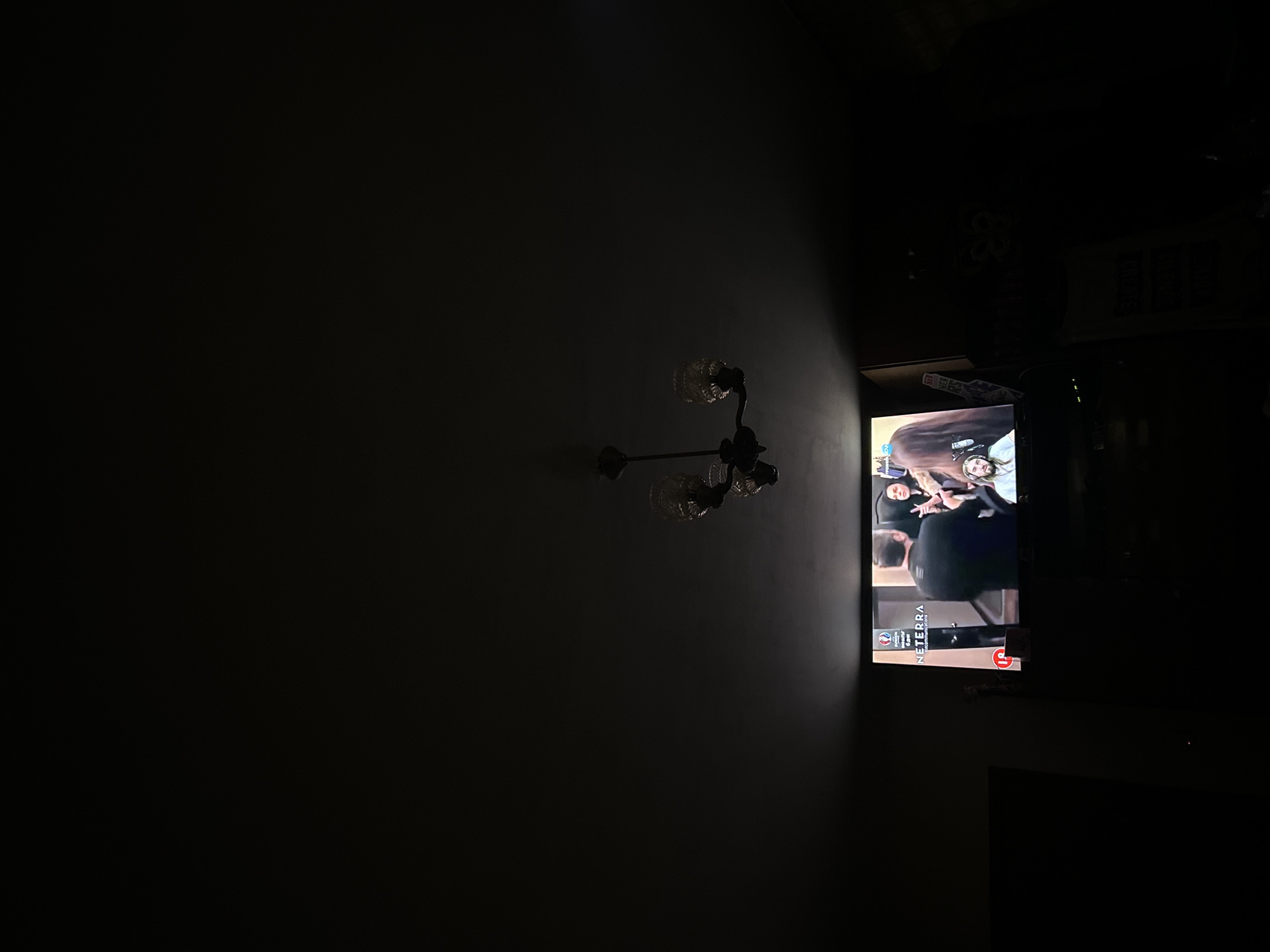 The image depicts a dimly lit room at night. A dark wall occupies the left side of the frame, with a decorative chandelier attached to it. To the right, a television screen illuminates a portion of the wall, displaying a scene with people. Details of the room are obscured by the darkness. There is no information to indicate the location is in Varna, Bulgaria.