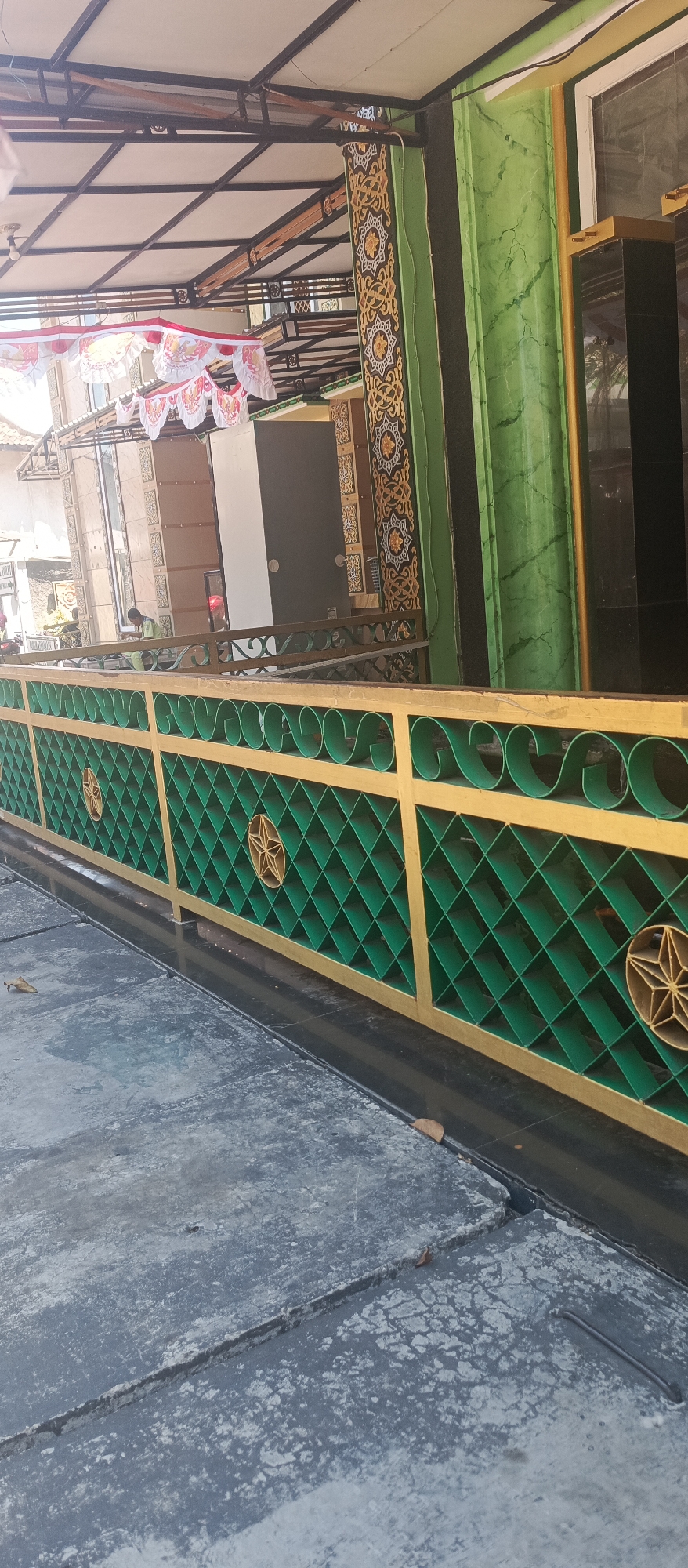 An exterior shot captures a decorative green and gold metal fence in front of a building. The fence features a diamond pattern with star embellishments. Behind the fence, a person wearing a light green shirt is visible, seated near a doorway. The building's facade is decorated with ornate golden and dark brown patterns on green columns. Festive red and white banners are strung across the overhead structure. The ground in the foreground is paved with rough, grey concrete slabs. The scene suggests a public or semi-public space, possibly a mosque or community building, in Situbondo, Indonesia, given the decorative elements and the presence of banners, which are common during national holidays.