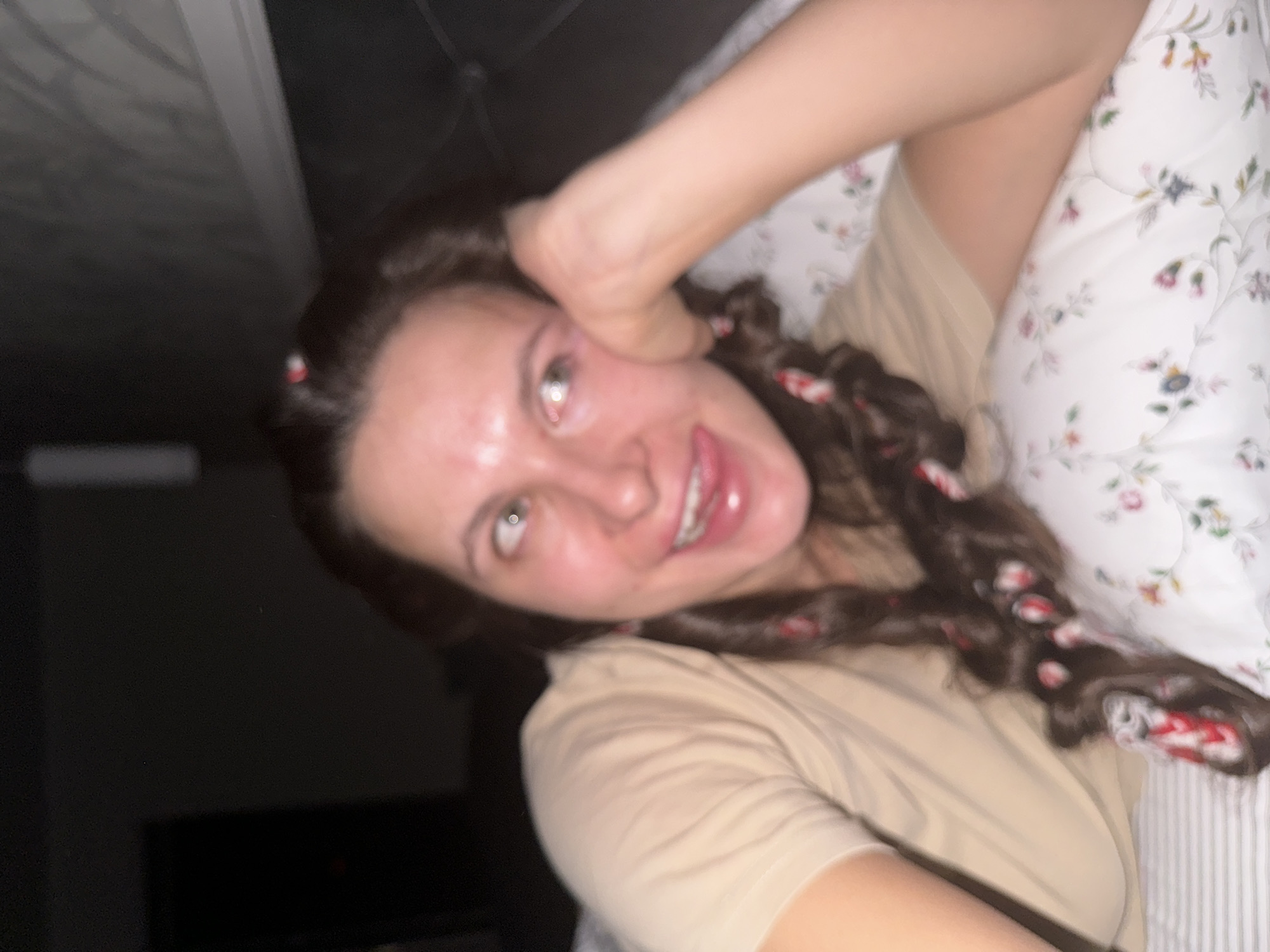 A woman with brown hair styled in pigtail braids, adorned with red and white ribbons, is lying on a floral-patterned pillow. She is wearing a tan shirt and appears to be playfully holding her hand up to her face. The background is dimly lit, with a dark object partially visible.