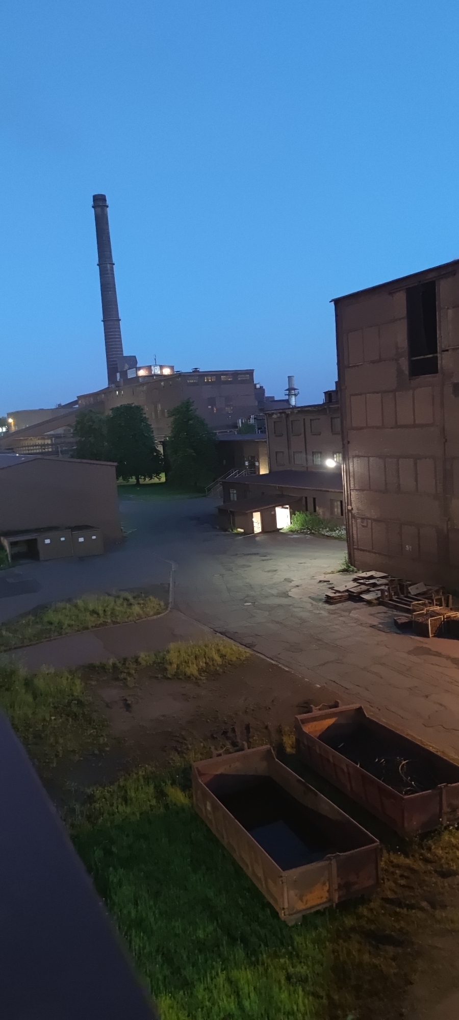 The image depicts an industrial area at dusk. In the background, a tall, slender industrial chimney pierces the clear blue sky. The chimney is part of a larger complex of industrial buildings, some of which are illuminated with warm light from windows, suggesting internal activity. In the foreground, there are several large, open-top industrial bins or dumpsters, some appearing empty and others containing dark materials, possibly waste or debris. The ground is a mix of paved areas and patches of overgrown grass. The scene conveys a sense of industrial activity during twilight hours, likely in an area of Salzgitter, Germany, known for its industrial heritage, particularly in steel production.