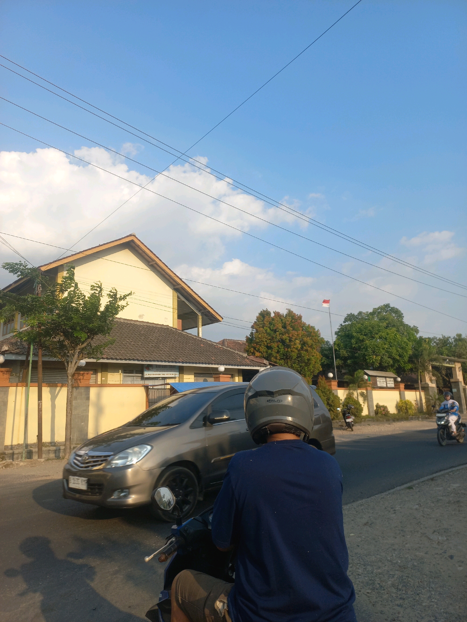 The image depicts a street scene in Baturetno, Indonesia. In the foreground, a person wearing a dark blue shirt and a grey helmet is seated on a motorcycle, viewed from behind. The motorcycle's handlebars and part of the seat are visible. To the left, the shadow of the motorcycle and rider is cast on the asphalt road.
In the middle ground, a dark grey car is driving on the road. The car appears to be a Toyota Innova, with its headlights and grille clearly visible. Further down the road, another motorcycle with a rider in a light blue and white uniform is approaching.
The background features a building with a light yellow facade and a brown tiled roof. Trees with green and reddish foliage are present near the building. A flagpole with the Indonesian flag (red and white) is also visible. The sky is blue with scattered white clouds. Electrical wires are strung across the sky. The overall environment suggests an urban or suburban setting during daylight hours, possibly late afternoon due to the angle of the sunlight casting shadows.