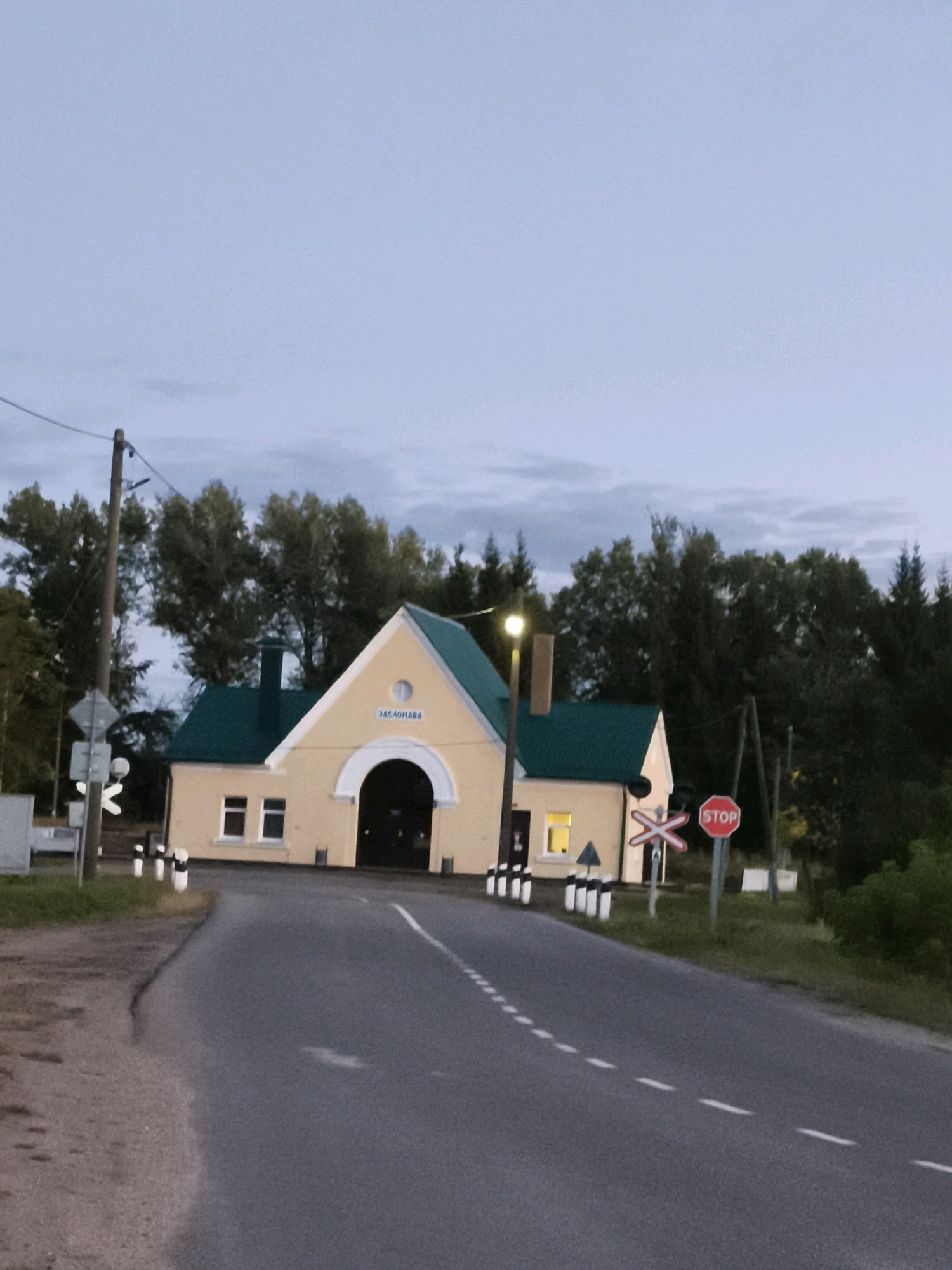 The image depicts a rural scene near the city of Gorski Sel'ski Sovet, Belarus, at dusk. A road curves towards a building with a light yellow facade and a green roof. This structure appears to be a railway station or crossing attendant's building, indicated by the inscription "ЗАГНАНАВА" above the arched entrance. There are windows and a chimney on the building. A lamppost with a light illuminates the area. Various road signs are visible, including a stop sign and a railway crossing sign. The road is paved and has white dashed lines marking the lanes. On the left side of the road, there's a dirt and grass verge, and to the right, more greenery and trees. The sky is overcast with a pale blue hue. There are no people visible in the image.
