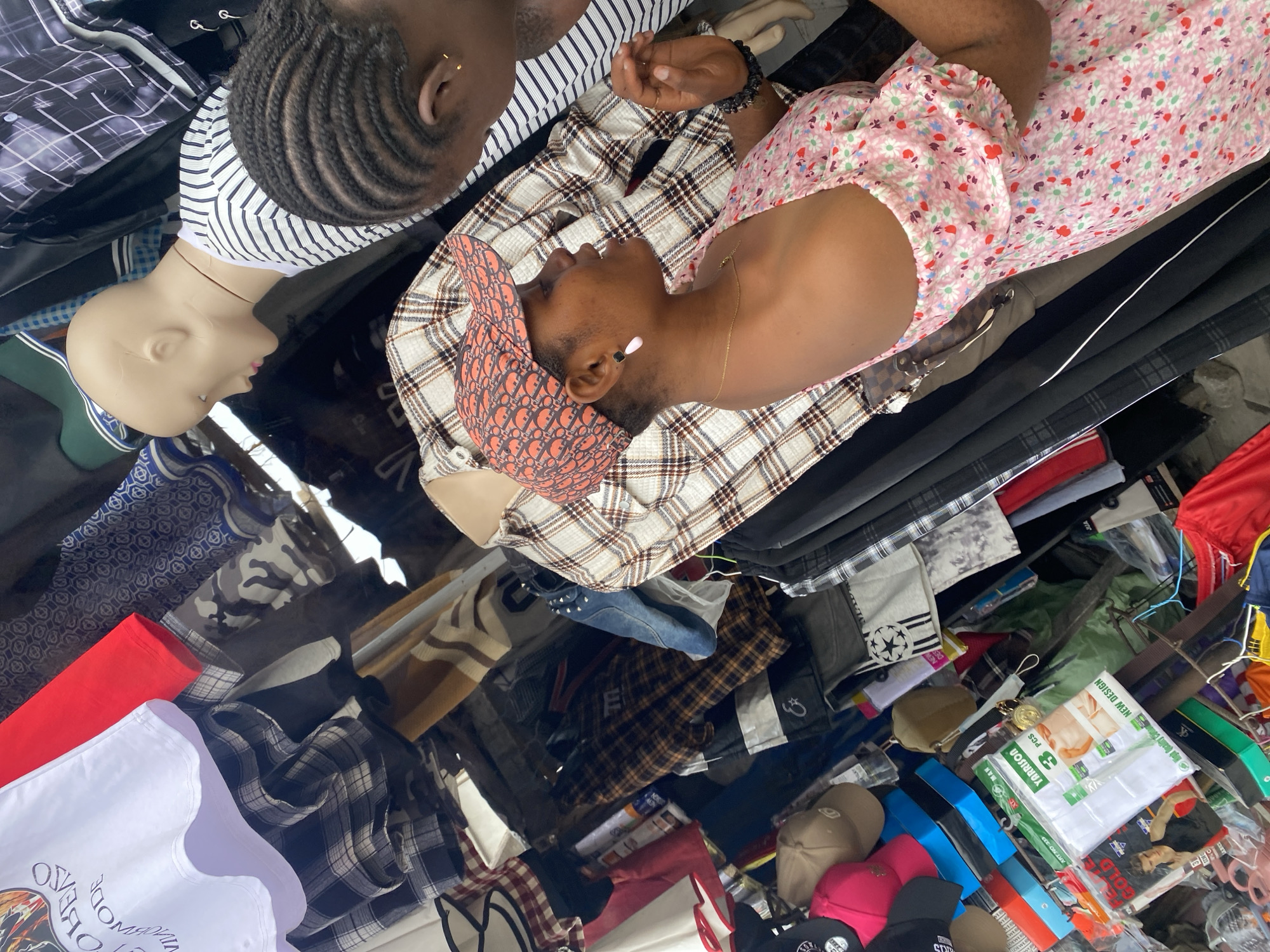 The image depicts a retail setting, likely a market stall in Lagos, Nigeria, showcasing a collection of clothing and accessories. Mannequins are present, one with braided hair wearing a striped top, and another bald-headed. A person wears a cap and is clad in a plaid shirt and floral dress. Garments of various patterns and colors surround them, including plaid shirts, t-shirts, and pants. Additional items such as hats, belts, and packaging materials are scattered throughout the scene.