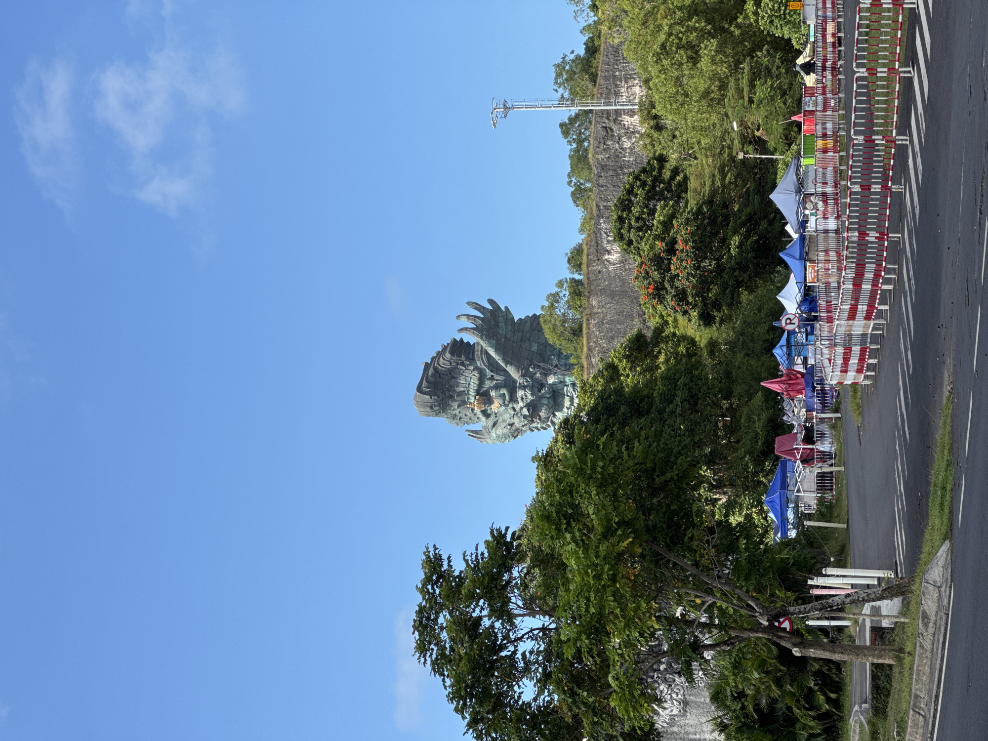 The image shows the Garuda Wisnu Kencana statue in Kuta Selatan, Indonesia, viewed from a low angle. The statue depicts the Hindu god Vishnu riding the mythical bird Garuda. It appears to be constructed of dark metal with intricate details visible on its wings and body.
The sky is clear and blue with faint clouds. In the background, there is a cliff face with trees and vegetation growing on top. A metal tower-like structure is also visible on the cliff.
On the right side of the image, there is a road with a row of tents or stalls along the edge, marked off by red and white barricades. There are a few visible people near the tents.
In the foreground, there are trees with green leaves and the side of what appears to be a structure or wall made of stone or concrete.