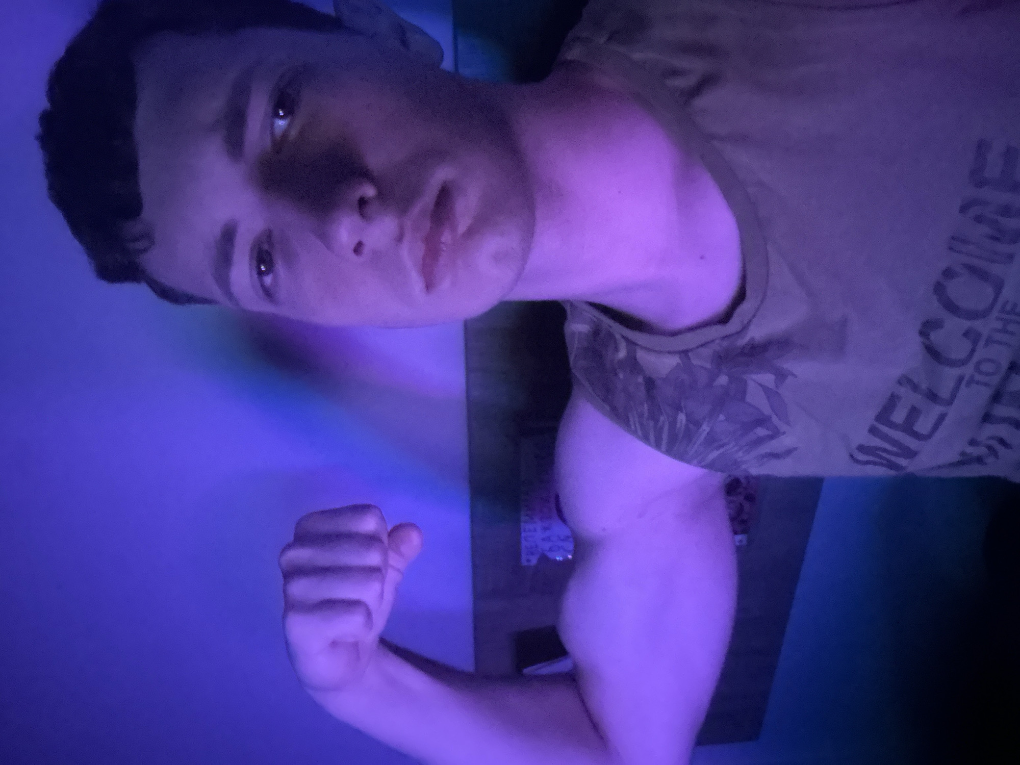 A young man is posing for a photo in a dimly lit room with purple lighting. He is flexing his bicep with his left arm, which is raised and bent at the elbow. His face is turned towards the camera, and he is looking directly at it. He has short, dark hair and is wearing a camouflage tank top. In the background, a blurred image on the wall appears to be a poster or a sign. The overall impression is that of a casual, self-taken photograph.