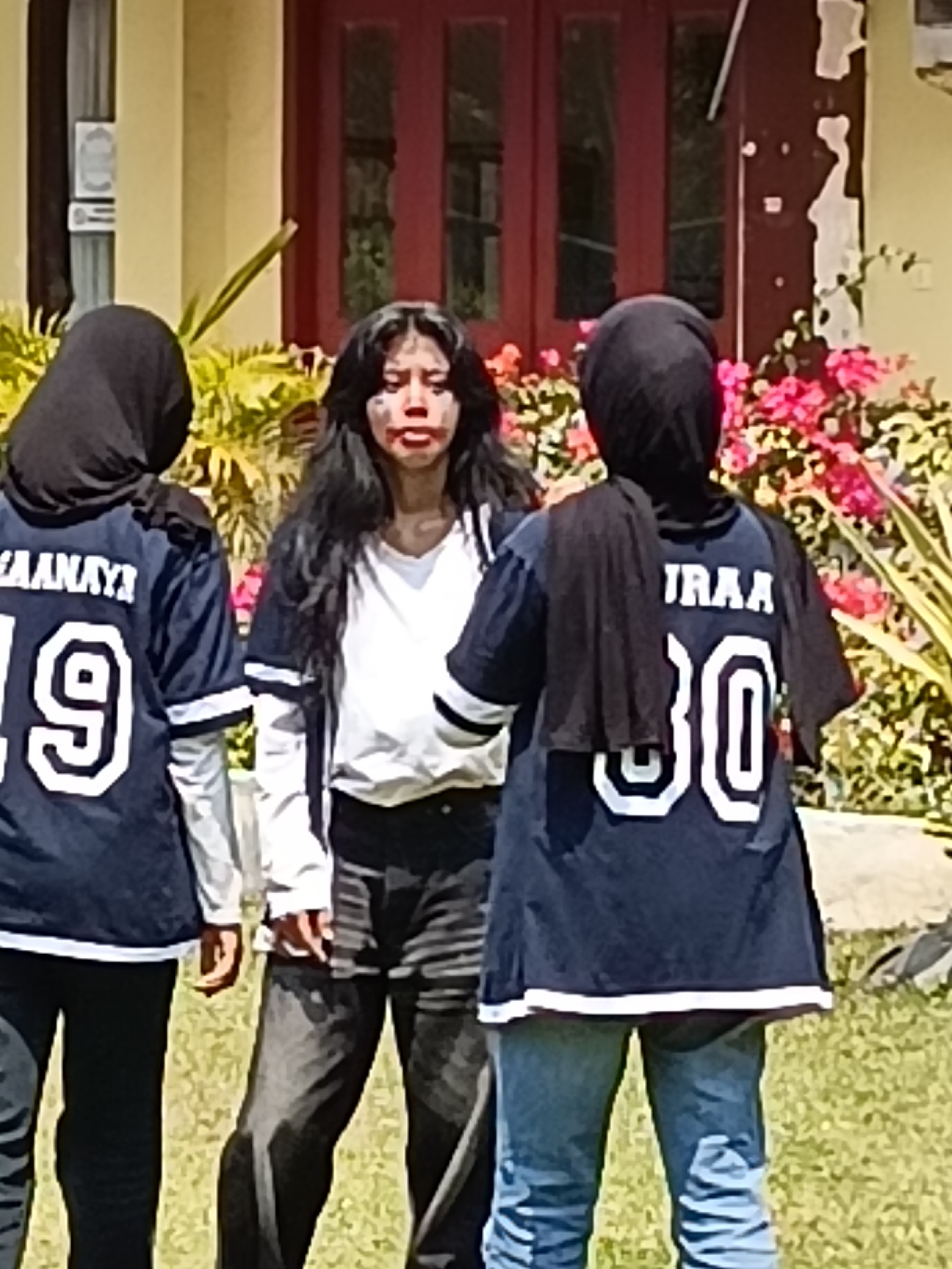 Three individuals, two wearing dark blue jerseys with white lettering and numbers, and one wearing a white t-shirt under a dark jacket, stand outdoors in front of a building with red doors and yellow walls. The individual in the center has dark hair and is wearing makeup on their face. The person on the left has their back to the camera, wearing a dark headscarf and a jersey with "AANAYAY" and the number "19" visible. The person on the right also wears a dark headscarf and a jersey with "IRAA" and the number "30". The ground appears to be covered in grass, and there are pink flowers in the background. The scene is likely a school or public area.