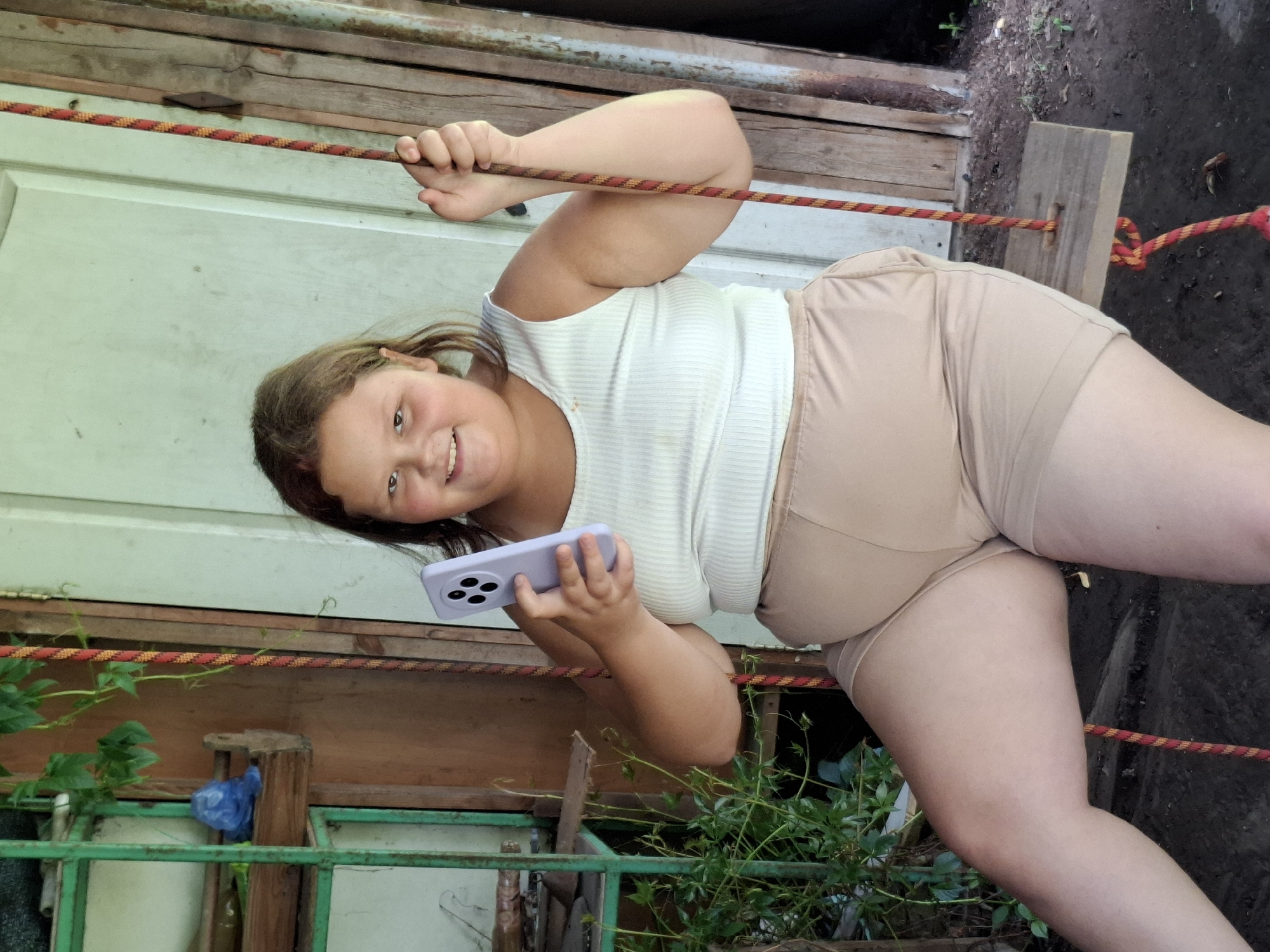 A young girl with light brown hair is holding a light purple smartphone in her right hand, looking at the camera and smiling. She is wearing a white, ribbed tank top and light brown shorts. The girl is standing, with her body tilted to her left, and her left arm is bent and resting on a red and yellow rope that extends horizontally across the frame. The rope appears to be part of a makeshift structure, possibly a playground or a practice area, as it's supported by wooden posts. The background consists of weathered wooden planks and a green metal railing with some greenery growing through it. The ground appears to be dirt or mud. The lighting suggests it's daytime, likely outdoors, but the specific location and weather are not discernible. There is no visible text in the image.