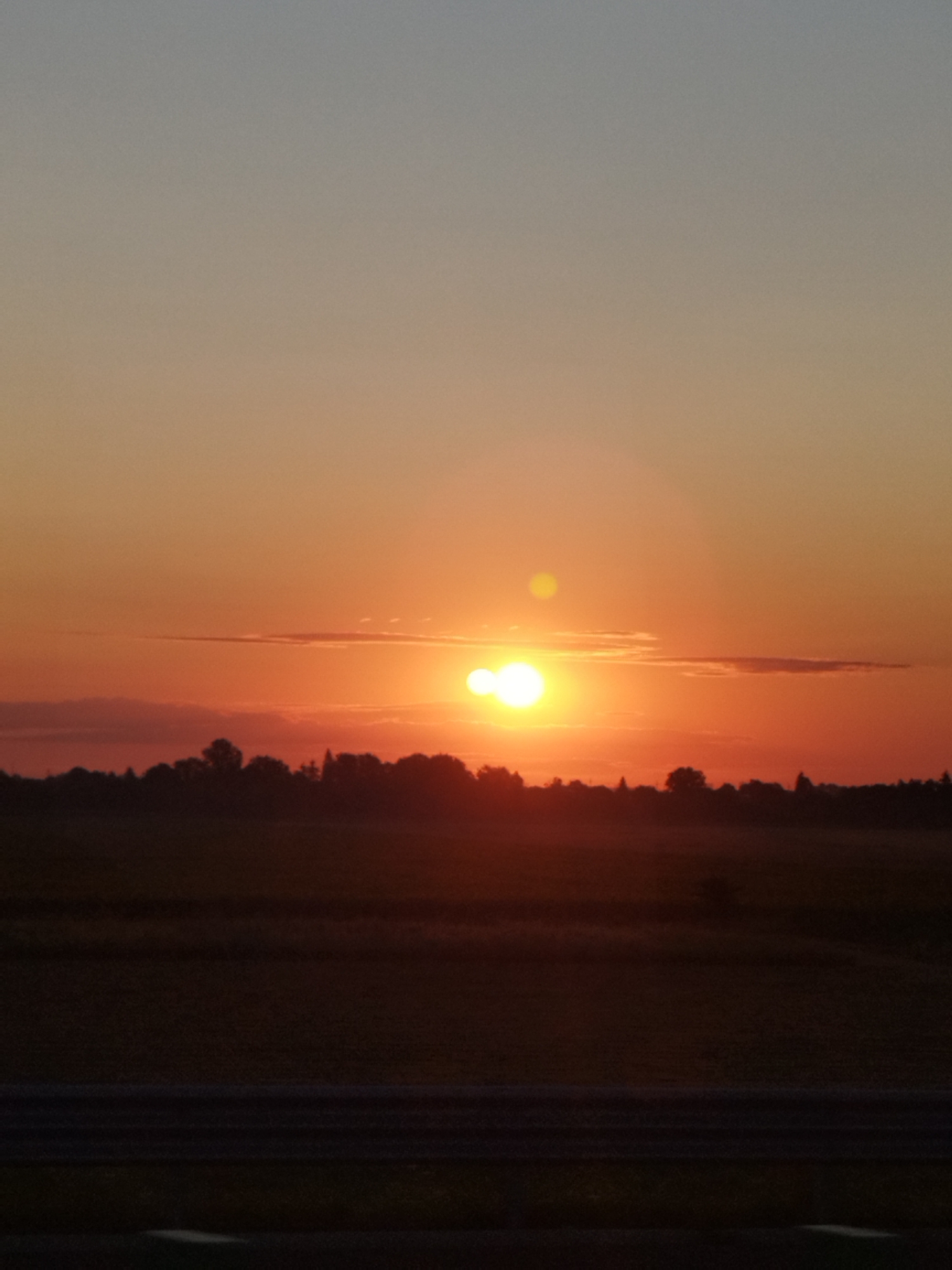 This is a vertical, full-frame, medium-distance, eye-level shot of a sunset over a rural landscape in Țegheș, Romania. The sky transitions from a pale gray at the top to a vibrant orange-red near the horizon. The sun appears as two bright, circular orbs, one larger and more prominent than the other, positioned slightly off-center to the left. Several thin, elongated clouds are visible in the sky. Below the sky is a dark, silhouetted tree line, indicating the edge of a wooded area. In front of the trees is a field, darkened by the low light, with subtle horizontal lines suggesting rows or textures. At the very bottom of the frame is a darker, horizontal band, likely a road or path, with faint horizontal striations. The overall scene evokes a peaceful, natural ambiance characteristic of a sunset in the Romanian countryside.