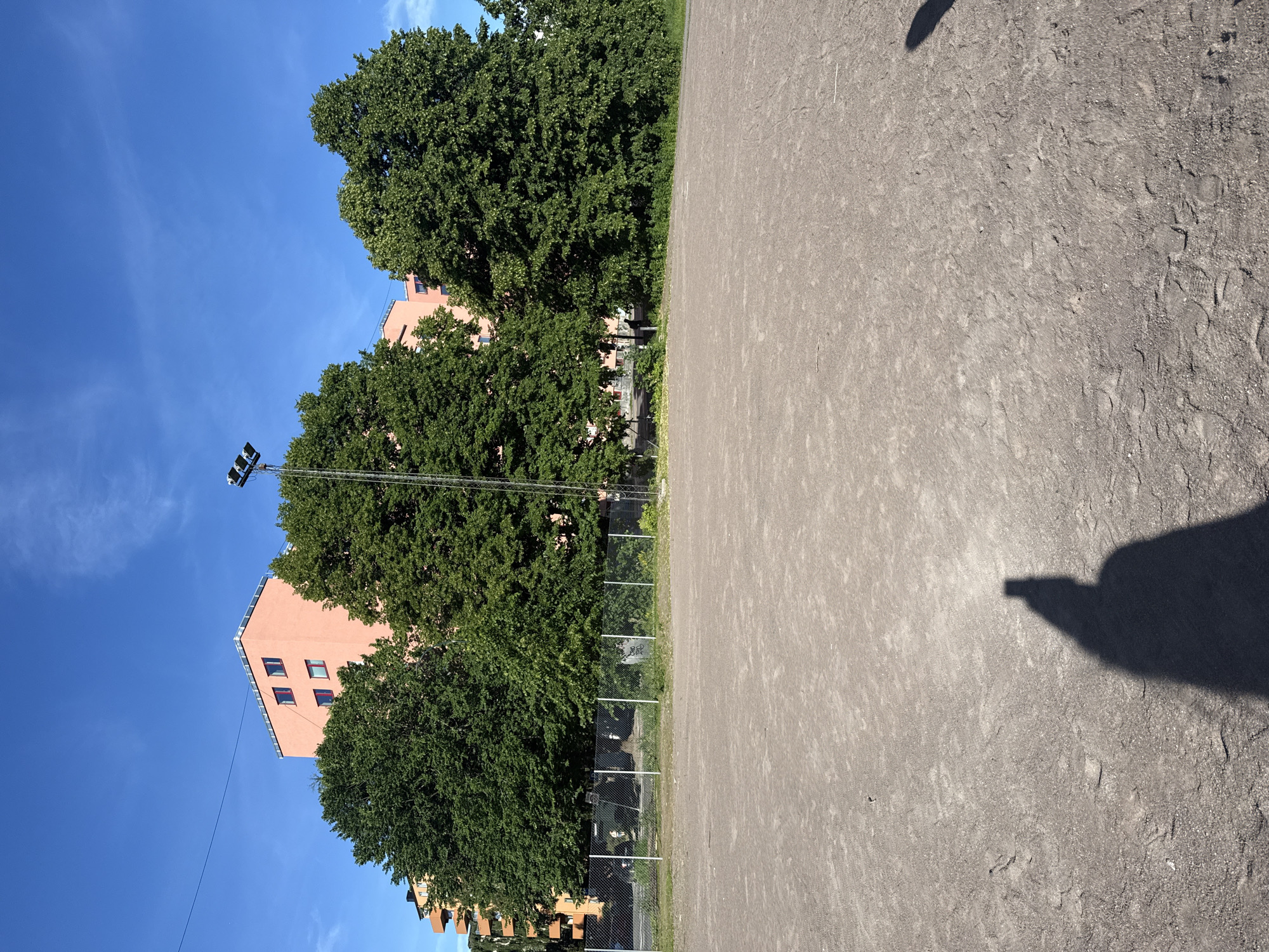 An outdoor scene on a clear, sunny day with a light breeze. In the foreground, a large, flat area of packed dirt or gravel stretches across the frame, with tire tracks and footprints visible. A shadow of a person holding a phone to take a picture is cast on the ground in the lower right corner. In the midground, a chain-link fence separates the dirt area from a line of mature, lush green trees. Behind the trees, a multi-story building with an orange-pink facade and red-trimmed windows stands against a bright blue sky. A tall metal pole with floodlights is positioned in front of the building and trees. A thin, dark electrical wire is strung across the upper portion of the frame. The setting appears to be an urban park or recreational area in Oslo, Norway.