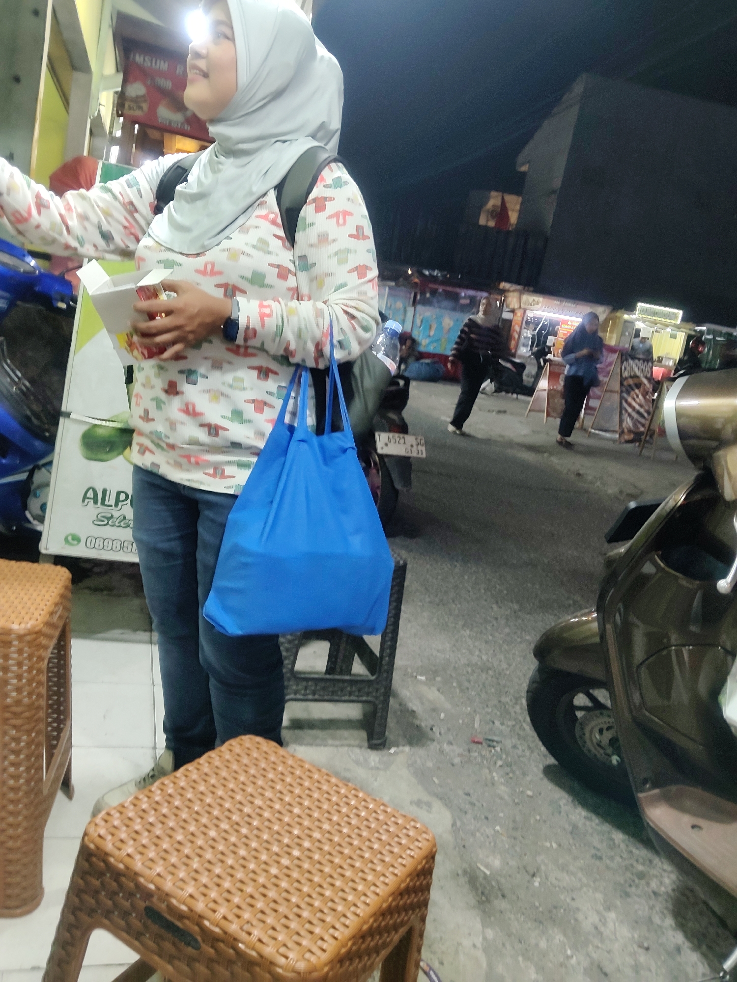 In Mekarmukti, Indonesia, a woman wearing a light grey hijab and a long-sleeved shirt with a repeating pattern of colorful clothing items stands outdoors at night. She is holding a white box in her hands, with her left hand inside the box. A bright blue tote bag hangs from her left shoulder. The woman is looking towards her left and smiling. She is wearing blue jeans and light-colored sneakers. In the foreground, to the left, is a brown wicker stool. Behind her, a light pole with bright lighting is visible. To her right, a brown scooter is parked. In the background, there are food stalls with illuminated signs and other people walking around. The ground is paved.