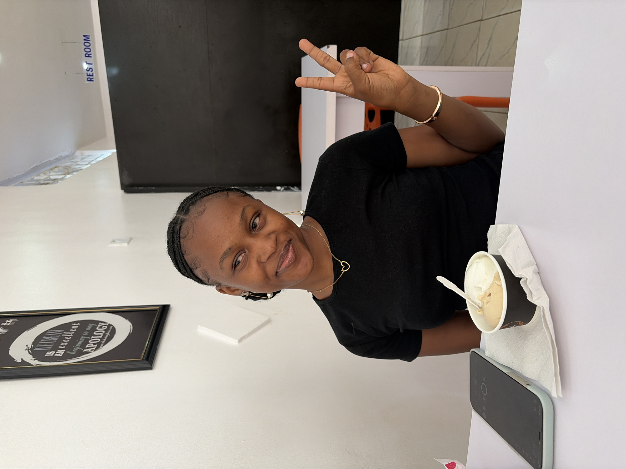 A young Black woman is captured in an indoor setting, likely an ice cream parlor or cafe, in Bar Jirgi Summa, Nigeria. She is smiling cheerfully and looking directly at the camera, posing with her right hand making a peace sign. She wears a black short-sleeved top, a gold necklace with a heart pendant, and gold earrings. Her hair is neatly braided.
To her right, on a white surface, sits a cup of ice cream with two distinct flavors (one lighter, one caramel-colored) and a white plastic spoon. Adjacent to the ice cream is a smartphone in a light blue case. The background features a stark white wall with a section of a dark cabinet or counter. A blue sign on the white wall reads "REST ROOM". On another wall to the left, a framed black and white artwork is visible, bearing the text "ICE CREAM IS AN EXCELLENT beginning to any APOLOGY". The overall atmosphere appears casual and joyful, suggesting she is enjoying a treat and a moment of leisure.