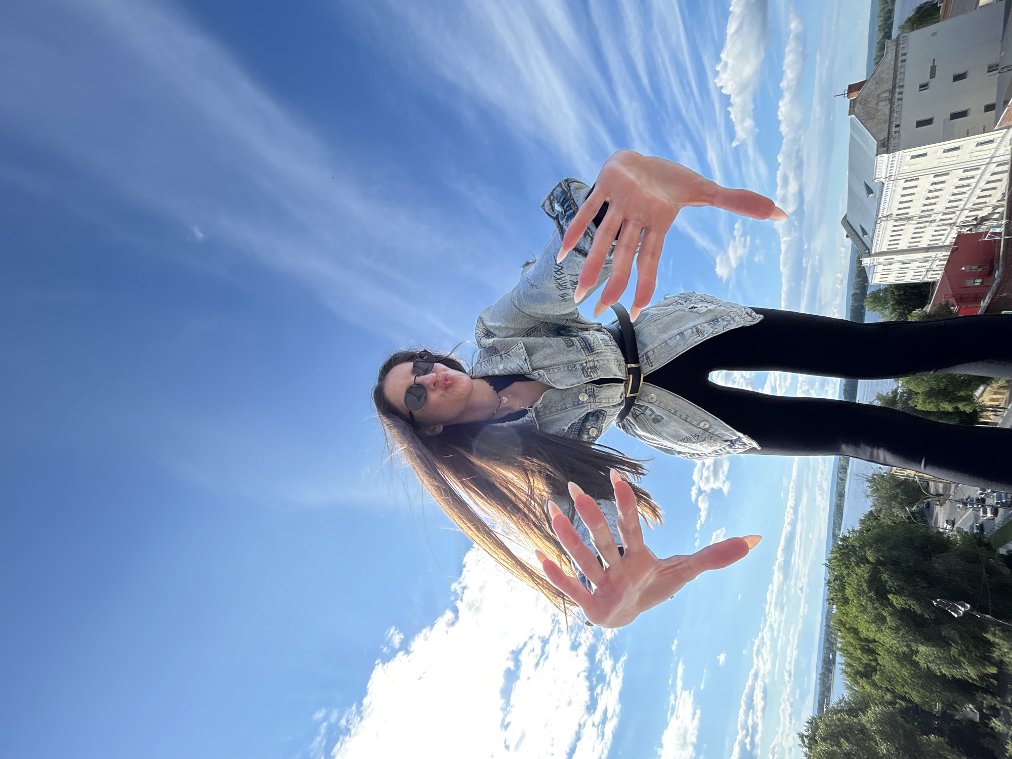 This is a low-angle, full-body, outdoor, eye-level shot of a young woman. She is wearing a denim jacket, black leggings, and sunglasses. Her hair is long and brown, and she has long, light-colored fingernails. She is posing with her hands outstretched towards the camera, with her face in a pouting expression.
The background is a bright blue sky with wispy white clouds. In the upper right portion of the image, there are buildings, a river, and green trees, suggesting an urban or semi-urban setting. The scene appears to be taking place during the daytime, under clear and sunny weather conditions.
The image is oriented sideways, with the woman appearing to be lying on her back or looking up from a low vantage point. There is no visible text in the image. The overall mood conveyed by the image is playful and casual. The location context indicates Novokuybyshevsk, Russia.