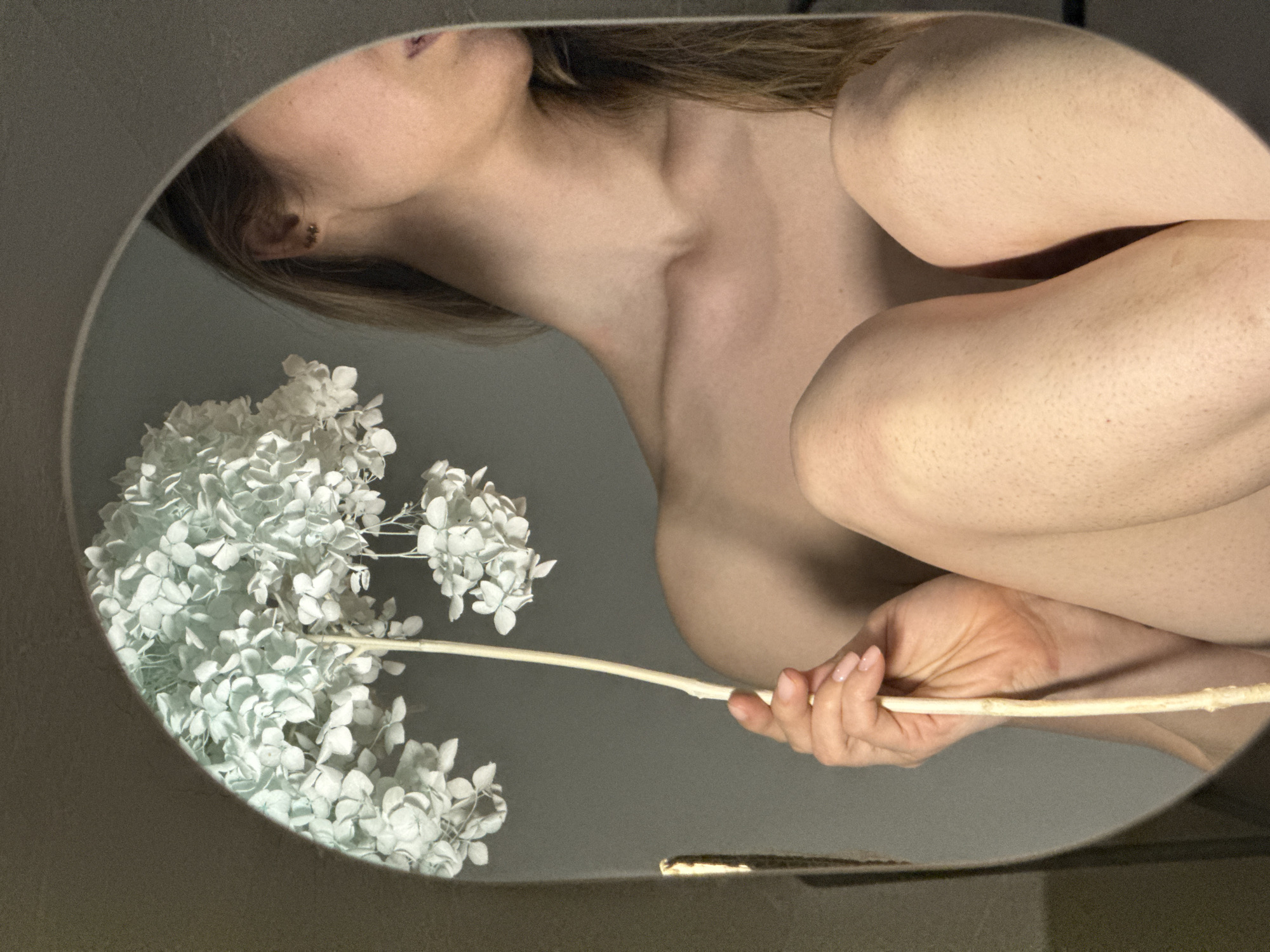 A partially nude person is holding a white flower, possibly a hydrangea, in front of them. The image is taken through a round mirror or similar reflective surface. The visible skin tone is fair. The person has medium-length brown hair. The background is a neutral gray. There are no indications of the city of Chaikovsky, Russia, or any other specific location.