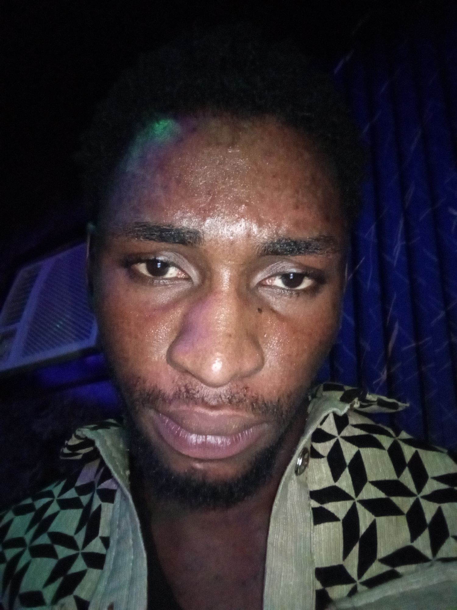 The image is a close-up, head-and-shoulders selfie of a Black male, appearing to be in his 20s, with dark skin and short, dark hair. He has sweat on his forehead and face. His eyes are open, and he is looking directly at the camera. He has a light beard and mustache. He is wearing a collared shirt with a black and light green geometric pattern.
The background consists of a blue, textured curtain on the right side of the image. On the left side, partially visible, is what appears to be the interior of an air conditioning unit with visible slats, and a faint colored light, possibly green, is illuminating the top of the man's head. The lighting conditions are dim, suggesting the photo was taken indoors at night or in low light.
The image does not provide any specific visual cues to indicate its location within the city of Lokoja, Nigeria.
