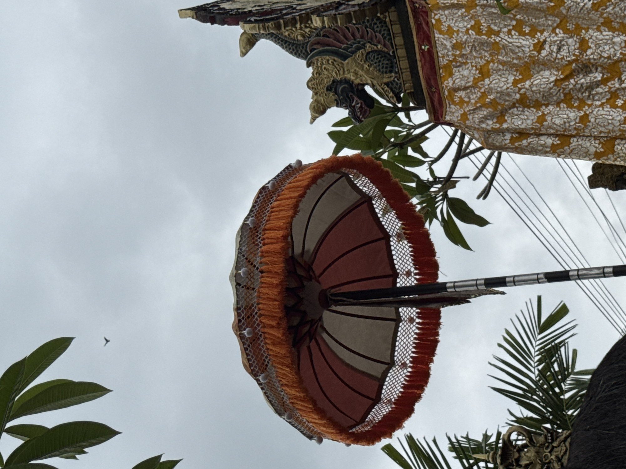 The image shows a low-angle view of decorative elements against a cloudy sky. A traditional Balinese umbrella, with orange fringe and intricate white netting, is prominently featured. Above it, a carved dragon head with gold and red accents adorns a structure, suggesting a temple or ceremonial site. A patterned yellow fabric with white floral designs hangs to the right, adding to the ornate aesthetic. Green leaves from nearby trees frame the scene, and a distant bird is visible in the sky. Power lines are also present, intersecting the sky and branches. The overall impression is one of cultural significance and artistry, likely observed during an outdoor event or festival. The cloudy sky suggests it might be overcast or a specific time of day where the light is diffused.