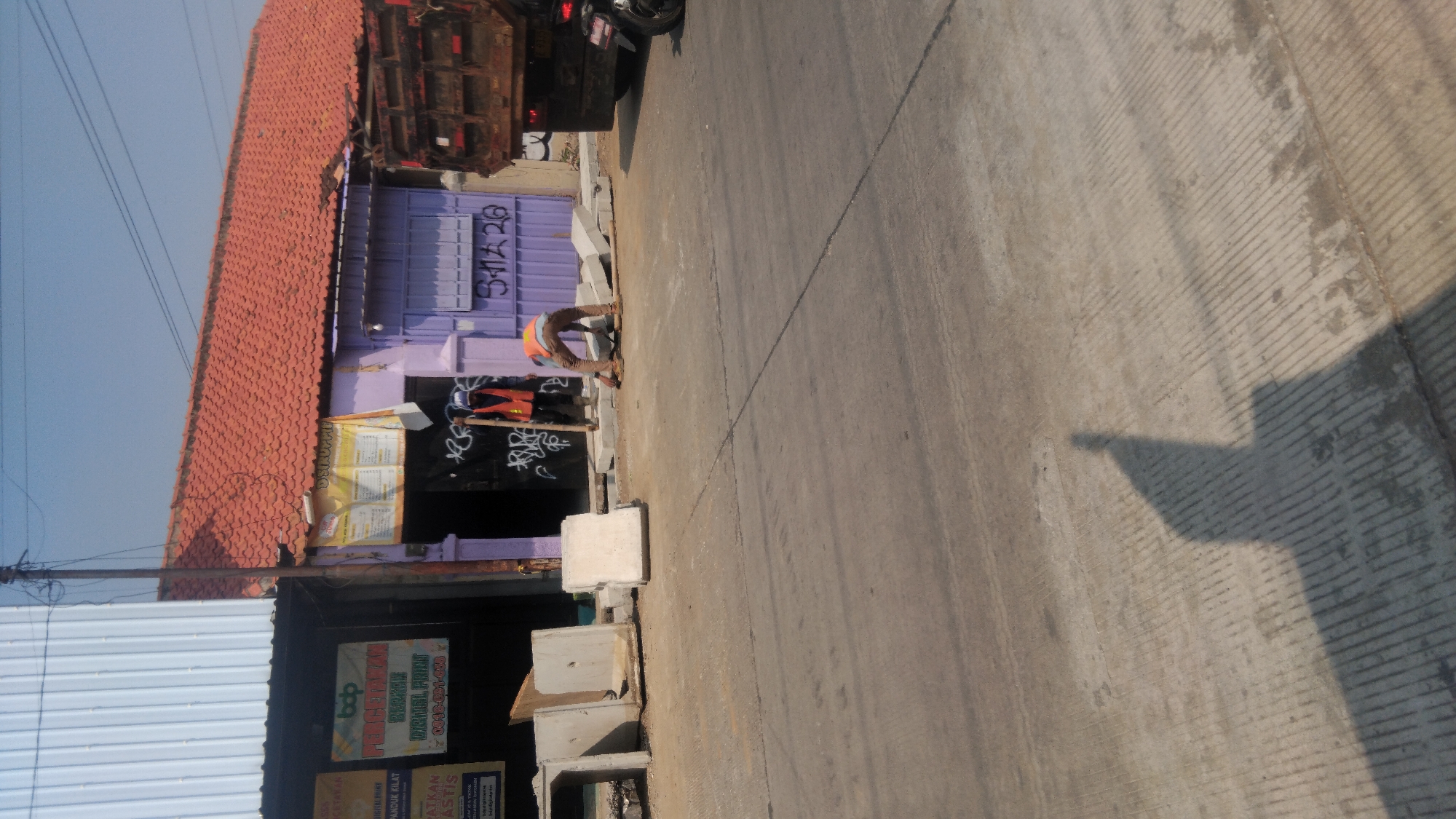In Batang, Indonesia, a street scene unfolds under a clear blue sky. A construction project is underway on the right side of the road, where two individuals in orange safety vests are working. One person stands, holding a wooden pole, while the other is bent over, appearing to be laying down concrete blocks. Several large precast concrete segments are stacked nearby, some on the side of the road and others in the process of being placed. A rusty red truck is parked on the right, its bed filled with materials. The ground is paved with asphalt and concrete, showing signs of wear and recent construction work. Buildings line the background, one with a red tiled roof and the other with a corrugated metal facade. Graffiti adorns the purple wall of one building, and a white banner hangs above an entrance. A motorcycle is visible parked behind the truck. The sun casts long shadows across the street, indicating it is likely late afternoon.