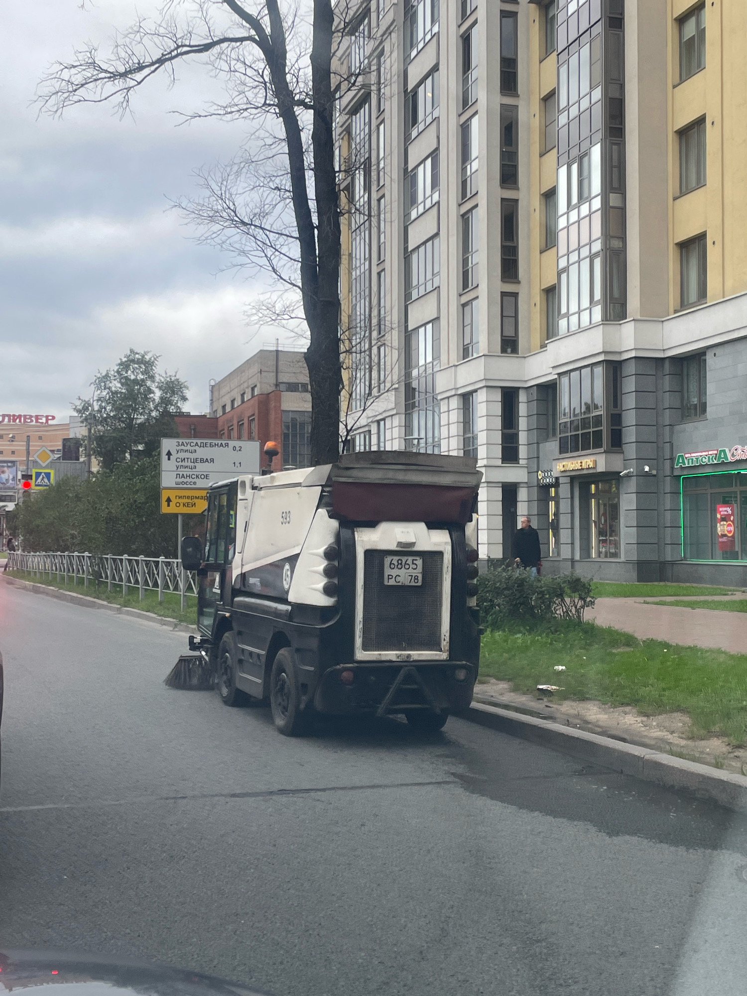 An outdoor, eye-level, medium shot captures a street cleaning vehicle in motion on a city street in Saint Petersburg, Russia. The vehicle, primarily white with some dark detailing and a visible license plate reading "6865 PC 78," is positioned in the right half of the frame, its brushes actively sweeping the asphalt. To the left of the street cleaner, a metal fence runs along the sidewalk, bordered by a lush green grassy area. In the background, a modern, multi-story building with a yellowish facade and numerous windows dominates the scene. A bare-branched tree is situated in front of this building, its branches reaching towards the overcast sky. Further in the background, various signs are visible, including directional signs in Russian. A person in dark clothing is walking near the building entrance. The overall atmosphere is overcast, suggesting a cool day.