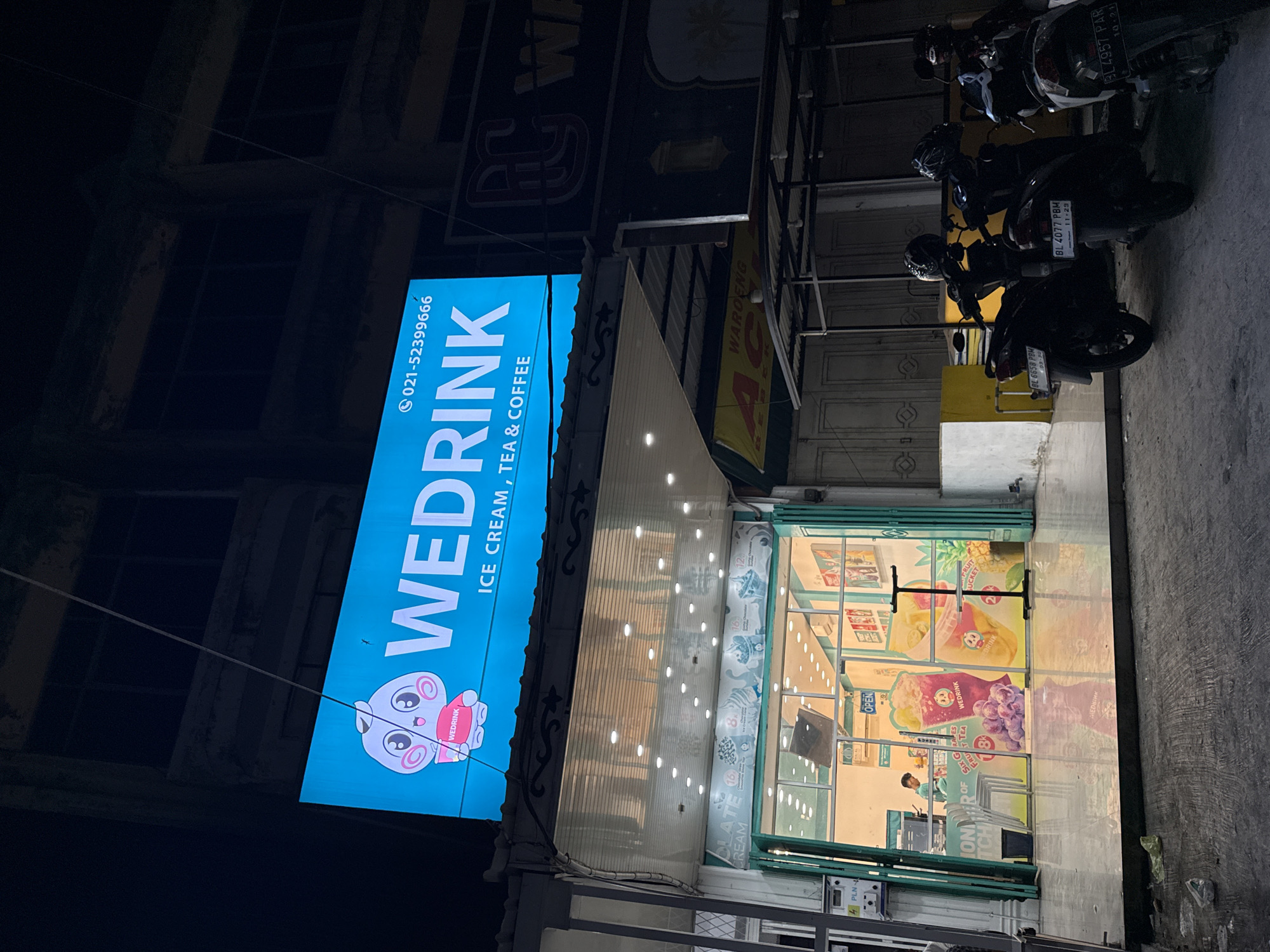 The image depicts the storefront of "WEDRINK", an ice cream, tea, and coffee shop. The store is in Sigli, Indonesia. The blue vertical sign displays the store's name, logo (a cartoon pig holding a "WEDRINK" cup), and the phone number "021-52399666". The shop's interior is visible through the glass doors and windows, with posters advertising the beverages and a worker standing behind the counter. Outside, three parked motorcycles are in the foreground. Above the storefront, there is a sign for "WAROENG".