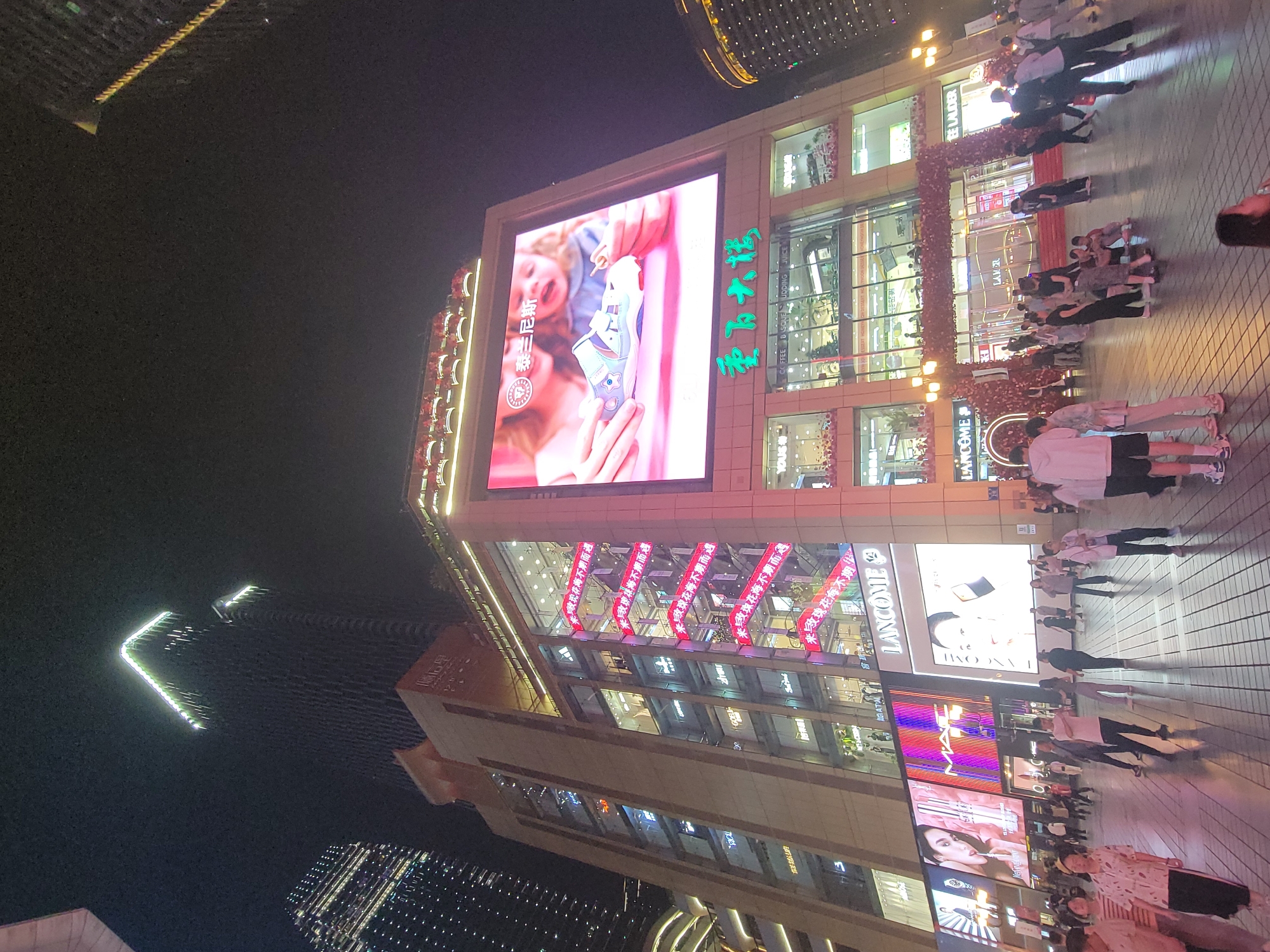 This media file captures a vibrant night scene in a bustling urban commercial area, identified as Yuzhong District, China. The central focus is a multi-story modern building, likely a department store or shopping mall, brightly illuminated and adorned with large digital screens and decorative lighting.
Numerous people are visible on the wide paved pedestrian area in front of the building. They appear to be walking, standing in groups, and engaged in evening activities typical of a busy city center. Some individuals are seen wearing casual attire. The ground is paved with a grid pattern, and reflections of the bright lights are visible on its surface.
Notable details include a large LED screen on the main building displaying an advertisement with a person holding a shoe. Another large screen shows beauty product advertisements, including "LANCOME." Several red banners with Chinese text hang from the building's facade, creating a festive atmosphere. A decorative archway or tunnel-like structure, covered in red floral or ribbon elements, frames an entrance. In the background, other tall, illuminated city buildings complete the urban skyline, with one distinct structure featuring a bright, angular "V" shape.
The time of day is clearly night, evidenced by the dark sky and the extensive use of artificial lighting from the buildings, screens, and streetlights. The scene suggests a lively and active commercial environment.
Visible text includes:
- "重百大楼" (Chongbai Department Store Building), a prominent name on the main building.
- "泰兰尼斯" (Talanis/Tyranis), likely a brand name, seen on the large LED screen.
- "玫瑰花海不期而遇" (A sea of roses unexpectedly encountered), repeated on red banners and possibly a screen.
- Brand names such as "LANCOME," "ESTEE LAUDER," "LAMER," and "MAC" are visible on various signs and screens.
- "UDDING JUICE" and "BEE COFFEE" are partially visible on storefronts.