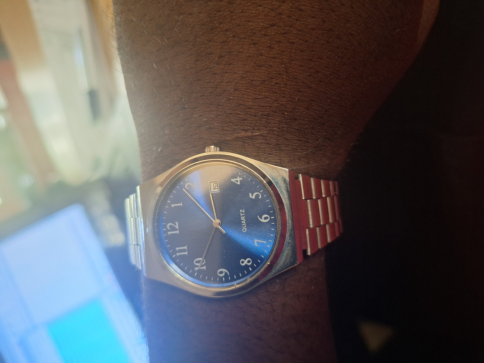 A close-up, eye-level, medium shot captures a man's wrist wearing a silver analog wristwatch with a dark blue face. The watch has silver hands, Arabic numerals, and a date window displaying the 19th. The watchband is also silver and made of segmented links. The man's skin, visible on his wrist and forearm, is dark brown and has visible hair. The background is blurred, showing a computer monitor in the upper left corner with a bright blue and white screen, and a lighter, out-of-focus background to the right, suggesting an indoor setting, possibly an office. The lighting appears to be artificial. No specific landmarks or details place this scene within Jalingo, Nigeria.