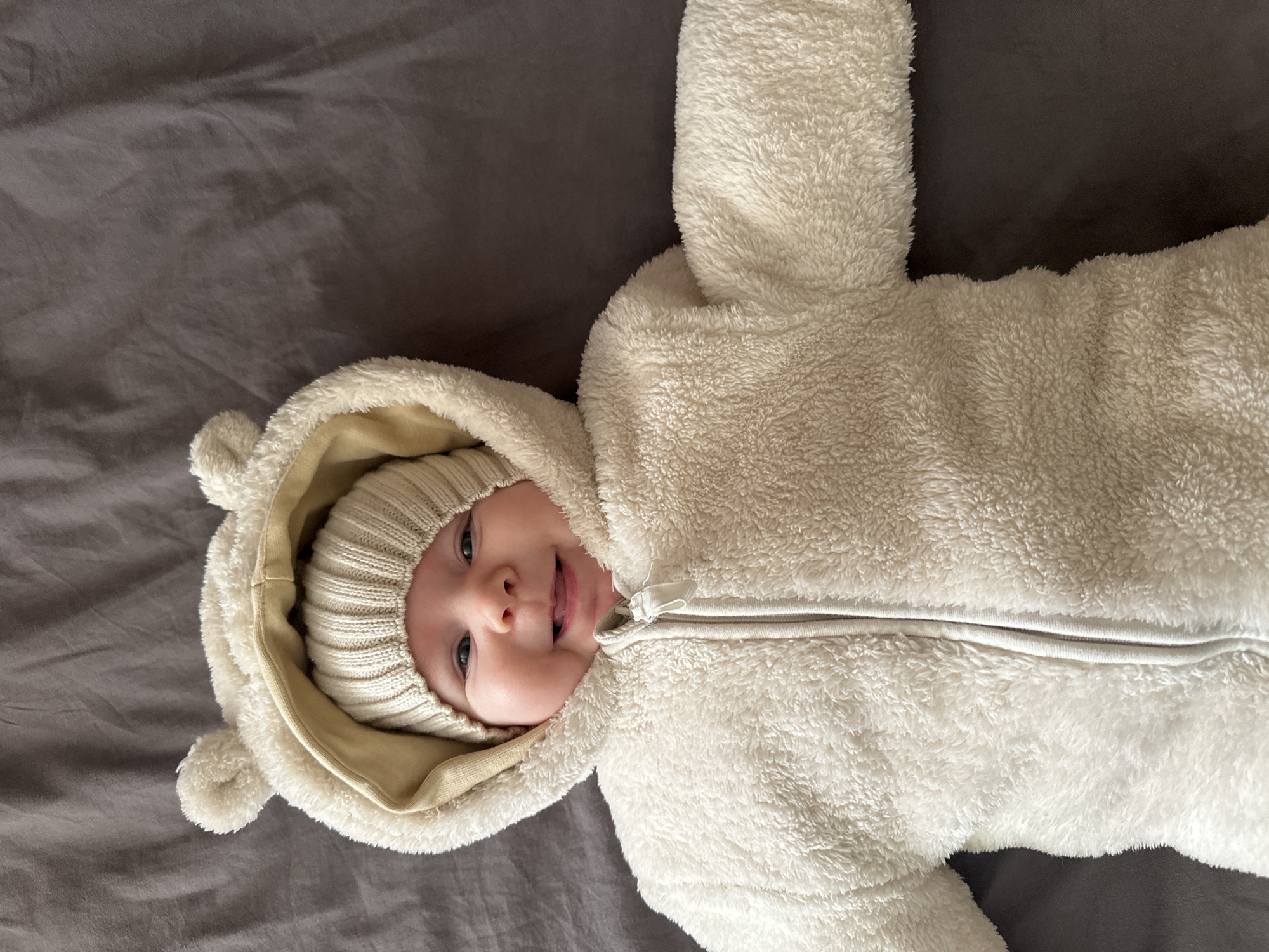A baby wearing a white furry jumpsuit with a hood is lying on a gray blanket. The baby's head is turned to the side, and the tongue is slightly sticking out. The hood has small bear-like ears on top. The baby is also wearing a knitted cap under the hood.
The location of the photo is impossible to define from the given image. There is no information available in the image that would associate it with a specific city.