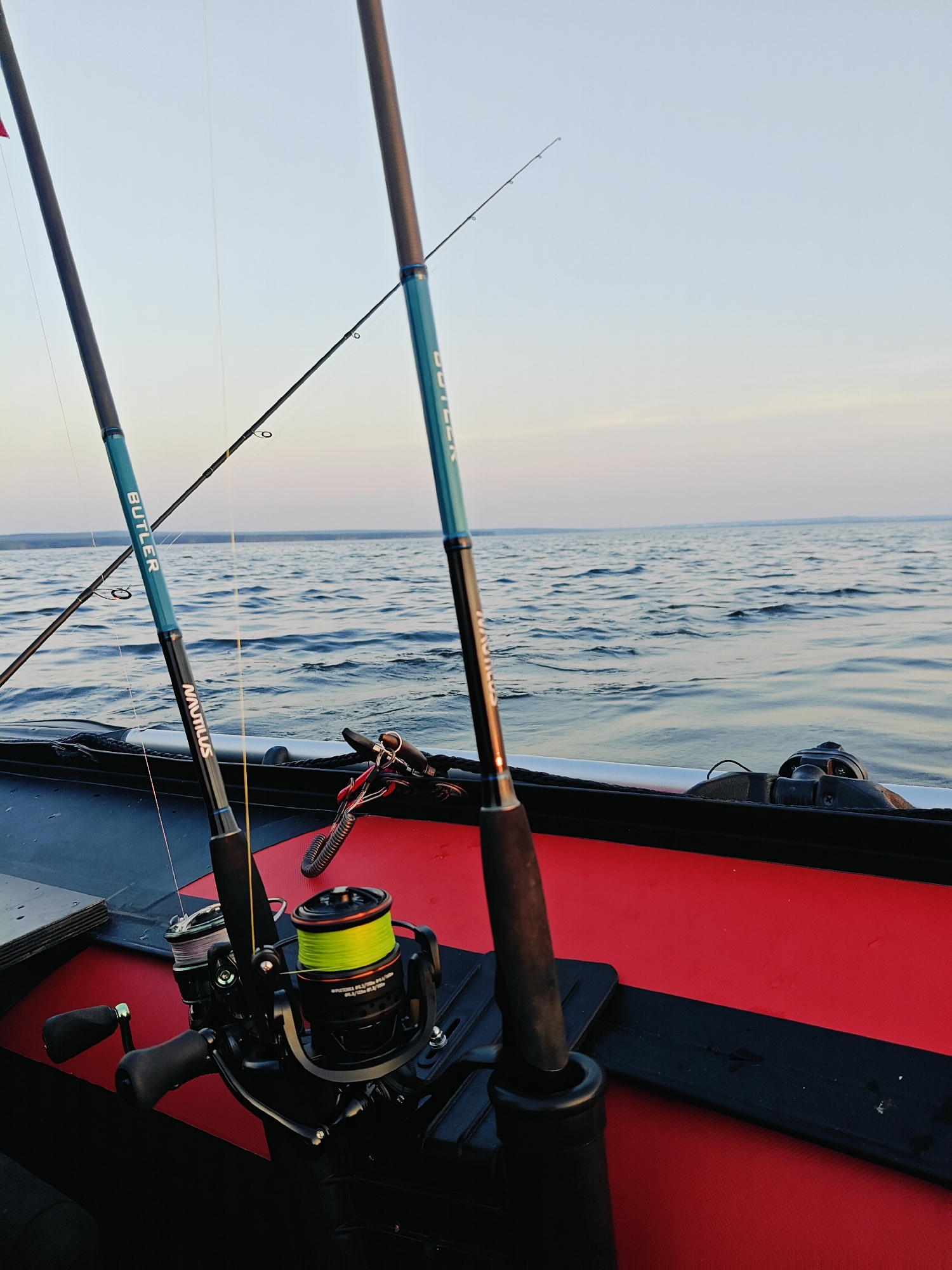 Two fishing rods are mounted on a red and black surface, likely the interior of a boat, with a body of water and a clear sky in the background. The fishing rods are dark gray with teal accents. One rod has the word "BUTLER" in white lettering on the teal section, and "NAUTILUS" in white lettering further down. A fishing reel with bright yellow fishing line is attached to this rod. The second rod, to the right, is partially visible and also has teal accents. Another fishing rod is visible further in the background, extending upwards and to the left. The water appears calm with gentle ripples. The sky is a pale blue with a hint of pink at the horizon. The scene suggests a fishing activity in an outdoor, aquatic environment. The location is identified as Chaykovsky, Russia.