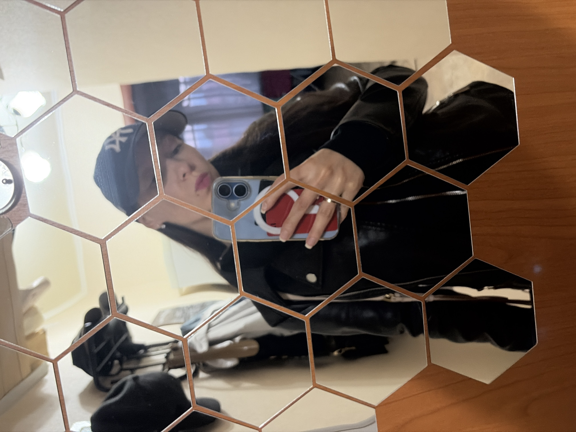 The image shows a reflection of a young woman in a hexagonal mirror. She is wearing a black leather jacket, a black baseball cap with a white "NY" logo, and a ring on her left hand. She is holding a smartphone with a red and white case in her right hand, taking a selfie. The background of the reflection shows a closet with clothes and hats, and a wall with light-colored paint. The mirror is mounted on a wooden surface. This scene appears to be an indoor setting, possibly a hallway or bedroom, and is not associated with the city of Япрыково, Russia.