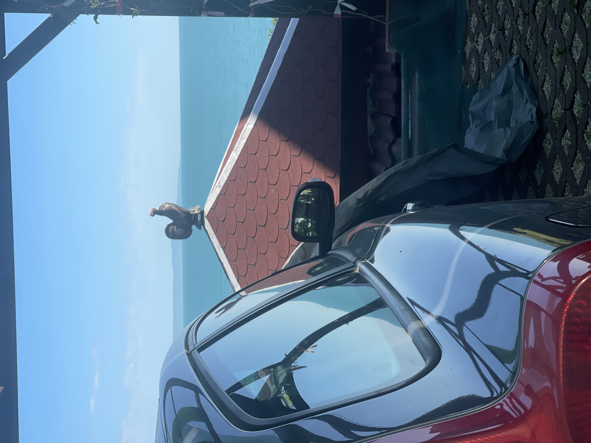 The image depicts a black car parked near a building with a reddish-brown roof with decorative roofing. In the background, there is a clear view of the sea and sky. The car's window reflects the surrounding environment. The location is likely Sozopol, Bulgaria, given the Mediterranean-style architecture and coastal setting. The overall scene is captured in bright daylight. The ground next to the building is paved with an open gridwork type paving.