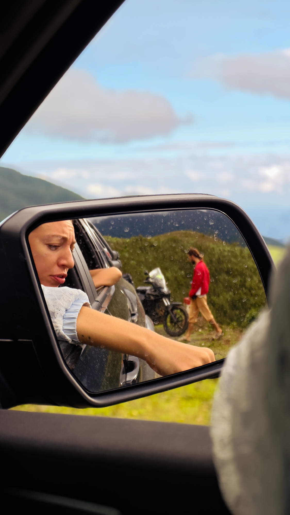 The image captures a scene from the interior of a vehicle, looking towards the passenger-side rearview mirror. The mirror's surface is speckled with numerous water droplets, indicating recent rain or mist.
Within the mirror's reflection, an adult woman is visible, leaning out the car window. She has light brown hair, and her expression suggests concern or contemplation, with her mouth slightly ajar and eyes directed downwards. She wears a light-colored, possibly white or pale blue, long-sleeved top with lace detailing. Her right arm, extended out of the window, exhibits several small, raised marks or scars.
Also reflected in the mirror, positioned outside the car on a dirt track, is an adult male. He has medium-length dark hair and a beard, wearing a bright red long-sleeved shirt and light-colored shorts or trousers. He is walking away from a dark-colored adventure-style motorcycle, which is partially obscured by his figure and features a light-colored front fairing. The man appears to be barefoot or wearing minimal footwear.
The background, both within the mirror's reflection and seen directly through the car window at the top, reveals a hilly or mountainous landscape characteristic of Xín Vàng, Vietnam. The terrain is covered in lush green vegetation, and the sky is a mix of blue with scattered white clouds. The immediate ground outside the car is vibrant green grass. The car's dark interior elements are visible in the foreground.