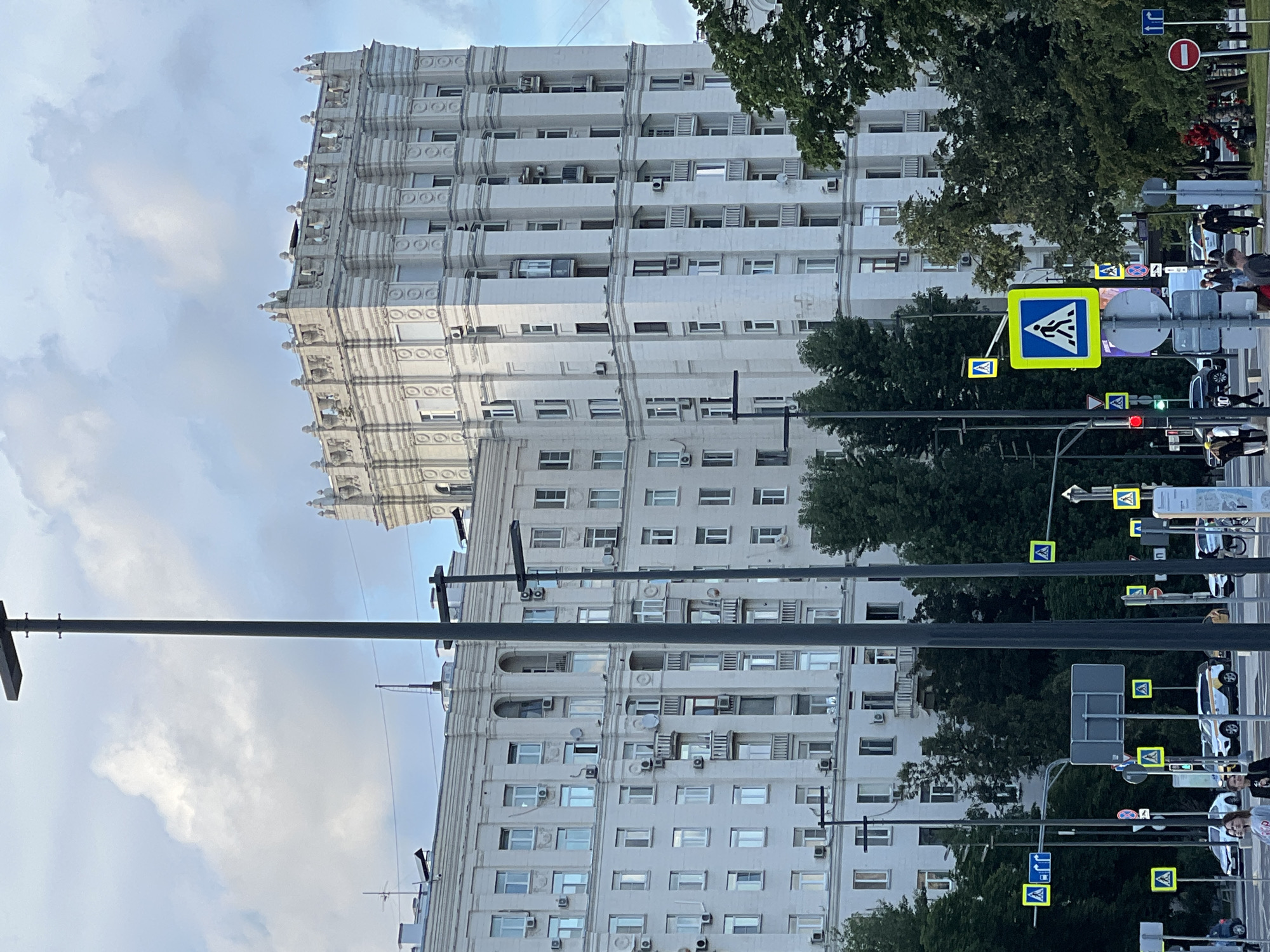 The image shows a street scene in Moscow, Russia. A large, white apartment building with ornate architectural details and many windows dominates the background. Trees line the street in front of the building. Several street signs and traffic lights are visible, including pedestrian crossing signs, a "no entry" sign, and a traffic light showing red and green signals. A few people are walking on the sidewalk and crossing the street, but their faces are not clearly visible. The sky is partly cloudy, suggesting it might be daytime. The overall impression is a typical urban street scene.