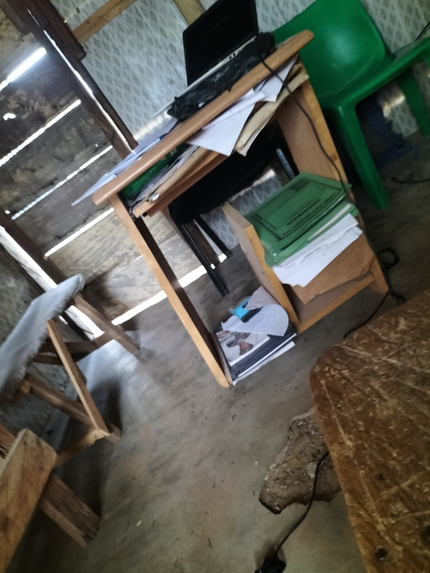 The image depicts an interior scene, likely a room with a rough, unfinished construction. In the foreground, a wooden table with a textured surface is visible, with a black cable extending from its vicinity. Behind it, a wooden desk is laden with stacks of papers and green folders. A laptop is open on the desk, and to the right, a bright green plastic chair is present. To the left of the desk, a wooden stool with a fabric seat is positioned. The walls and ceiling appear to be constructed from unfinished wood and corrugated metal, with light filtering in through gaps. The floor is concrete. The overall impression is one of a simple, functional workspace, possibly in a less developed area. The scene is consistent with a dwelling or small office space in Jalingo, Nigeria, characterized by its modest construction and the presence of common household and office items.