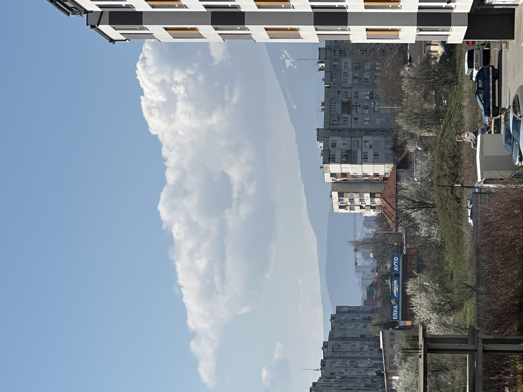 The image captures an urban scene in Sofia, Bulgaria. In the foreground, sections of two buildings are visible, one with a distinctive pattern of alternating dark and light horizontal stripes. Beyond the buildings, a cityscape unfolds featuring residential blocks of varying heights and designs. A prominent cloud formation dominates a significant portion of the frame, possibly obscuring distant landmarks. A range of vehicles are parked along a street near a grassy slope. Billboards with Cyrillic lettering are present, including the word "АУТО" (AVTO). The scene is captured under a bright, daytime sky, offering clear visibility.
