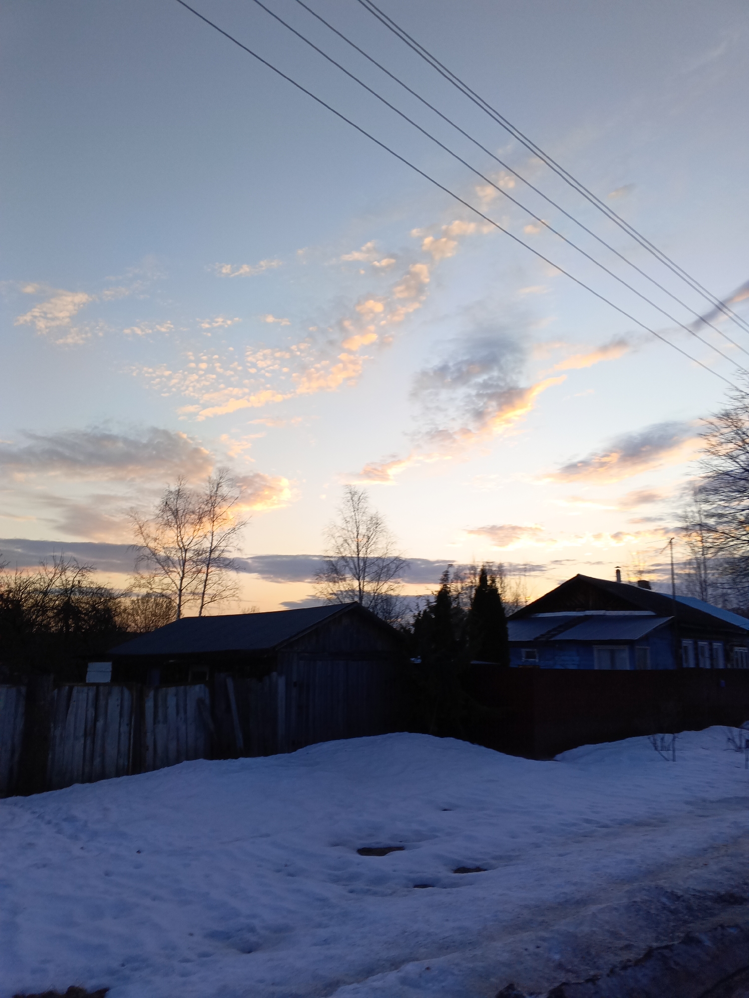 The image depicts a rural or semi-rural winter scene at sunset in Красный Маяк, Russia. The upper two-thirds of the frame are dominated by a sky exhibiting a gradient from light blue at the zenith to an orange-yellow glow near the horizon, with scattered stratocumulus and cirrus clouds illuminated in pink, orange, and grey hues. Four parallel dark utility lines traverse diagonally from the upper right toward the mid-left sky. The mid-ground features a collection of silhouetted bare deciduous trees, primarily on the left and center, and a darker cluster of coniferous trees on the right. Below the trees, a dark, single-story wooden outbuilding or shed with a pitched roof is visible behind a weathered wooden fence on the left. To the right, a two-story house, painted light blue, with a dark gable roof and a visible chimney, features multiple windows on its illuminated side. The foreground consists of an uneven expanse of snow, exhibiting a bluish tint from the low evening light, with darker, possibly wet or muddy ground visible at the bottom right edge. There are no visible people or discernible actions.