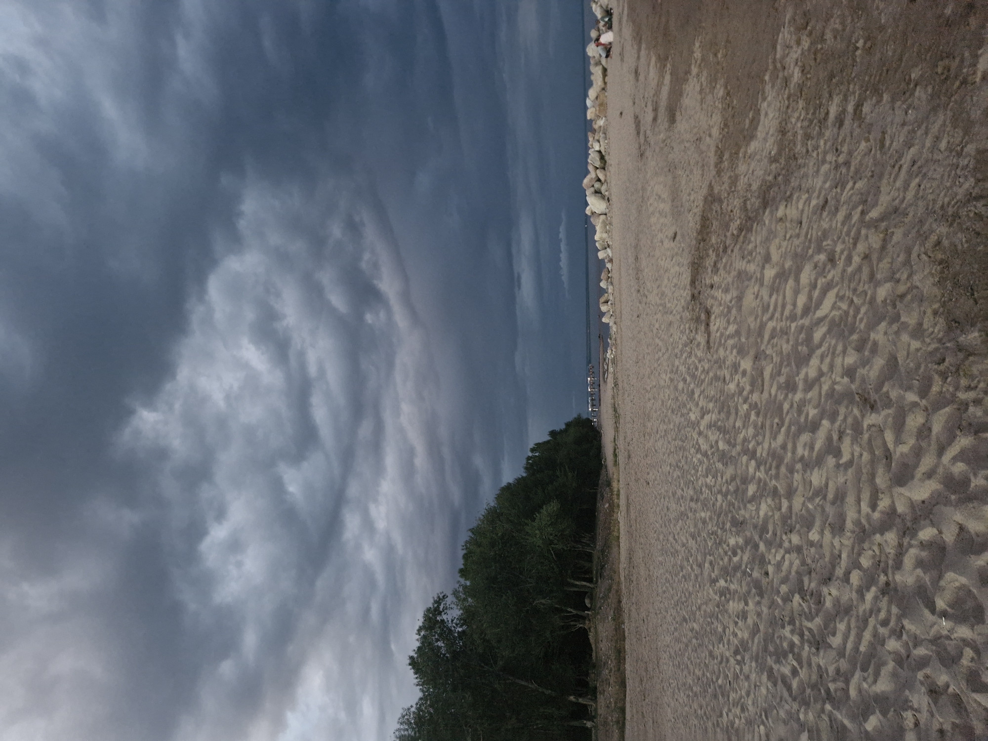 A wide-angle, vertical shot captures a beach under a stormy, overcast sky. The foreground is dominated by sand with subtle ripples and indentations. To the left, a dense line of dark green trees forms a natural border. Beyond the trees, a body of water is visible, and a pier stretches out into it. Along the right side of the beach, a line of large, light-colored rocks serves as a barrier. Two small figures, likely people, are seated on the rocks in the far right of the image. The overall atmosphere is somber due to the dark, dramatic clouds. The location is identified as Bulan-Segettu, Kyrgyzstan.