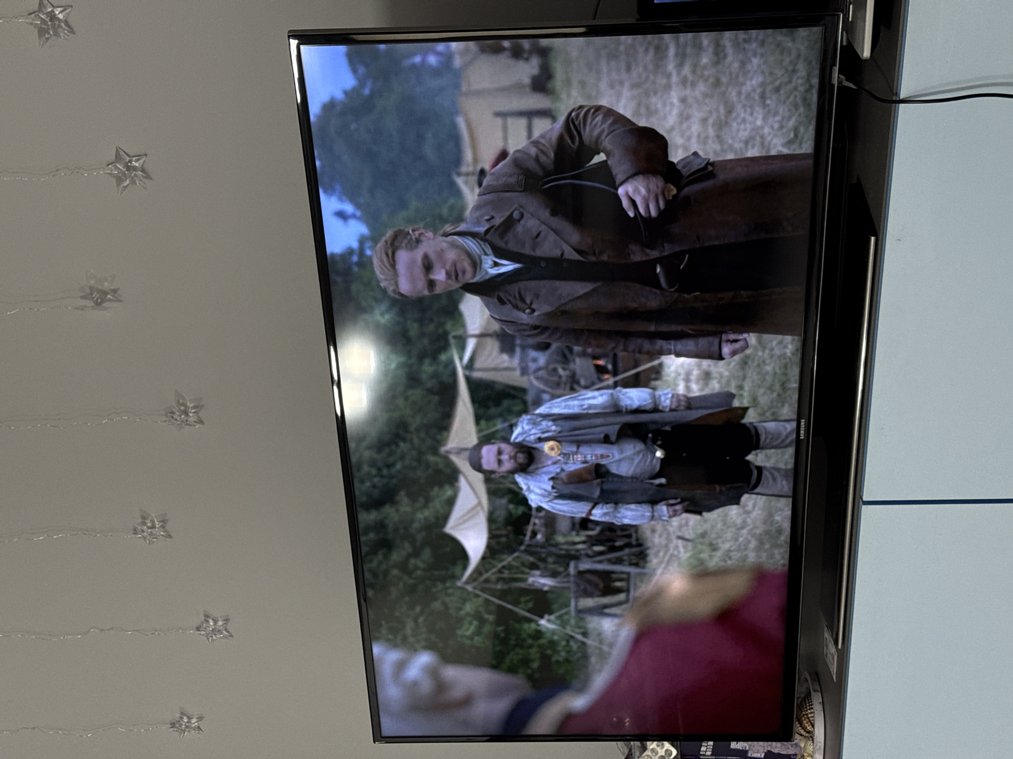 A Samsung television screen displays a scene from a historical drama. Two men are visible in the foreground, with a camp setting and trees in the background. The man on the left is wearing a red uniform with gold trim, his back is to the camera. The man on the right is wearing a brown leather coat over a white shirt and vest. He has his hair pulled back and is holding a dark object in his right hand. Behind them, tents are set up, suggesting a military encampment. The overall lighting indicates it is daytime, possibly late afternoon. The television is placed on a light blue cabinet. On the wall above the television, several clear star-shaped ornaments hang from thin strings. This image was taken in Łódź, Poland.