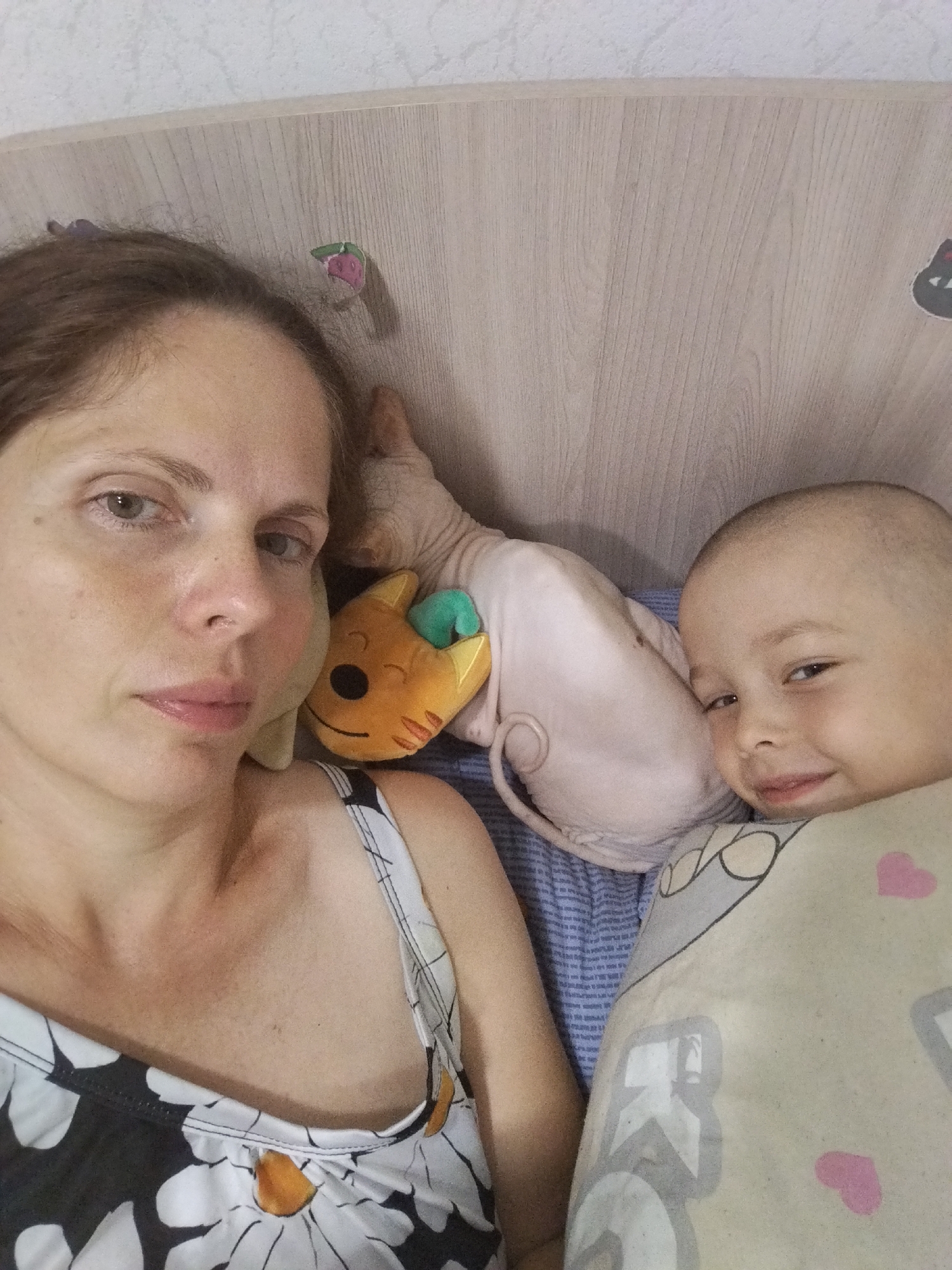 A woman and a child are lying in bed. The woman is on the left, looking at the camera with a neutral expression. She has brown hair and is wearing a black and white floral tank top. The child is on the right, looking at the camera and smiling. The child is bald and appears to be wrapped in a patterned blanket. Between the woman and the child is a pink hairless cat, also known as a Sphynx. The cat is lying down. In front of the cat is a small, stuffed toy that resembles a cat with green ears. The background appears to be a wooden headboard. The overall environment suggests a domestic setting, possibly a bedroom. The image was taken in Zavodoukovsk, Russia.