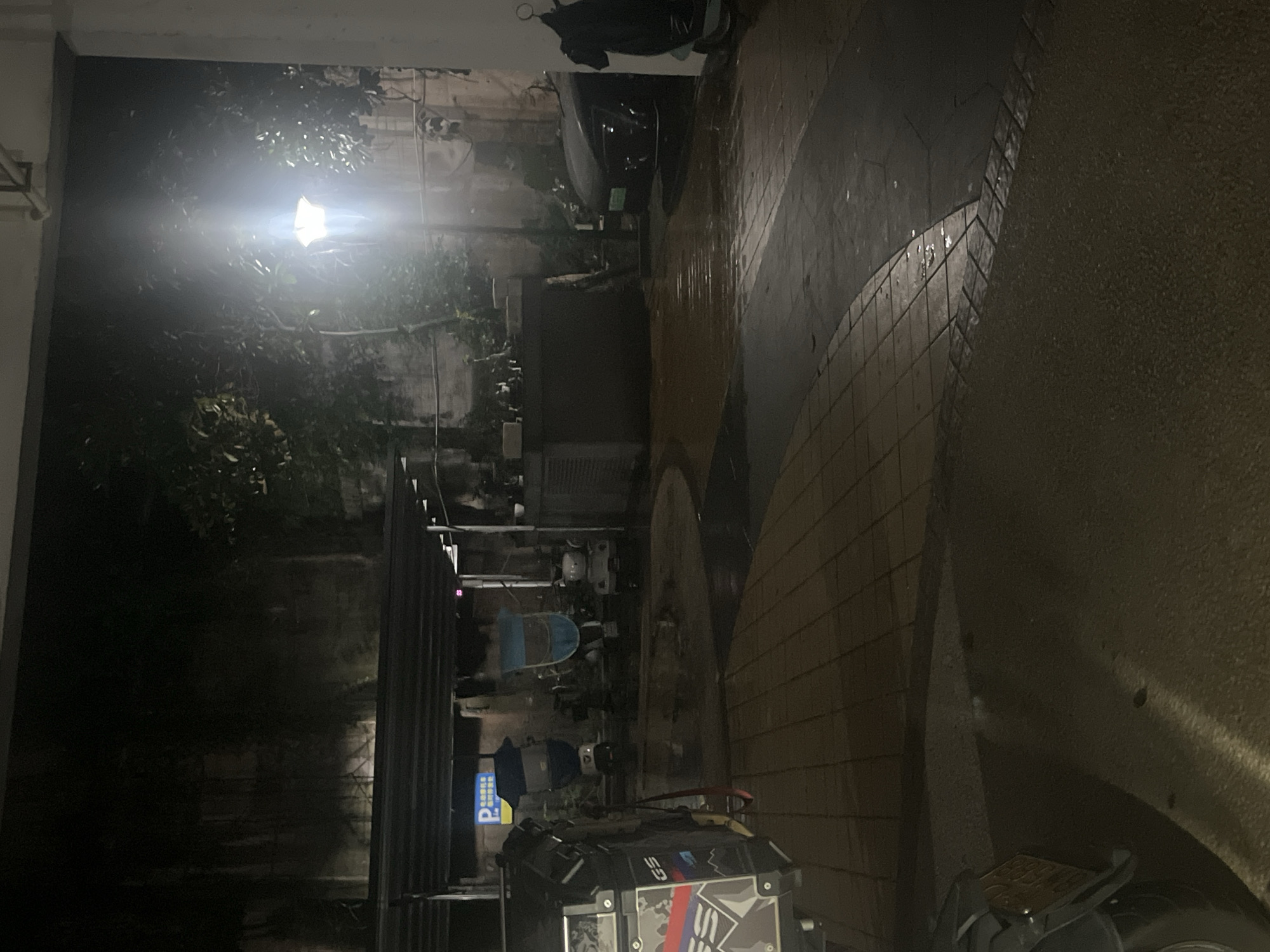 The scene depicts an outdoor, dimly lit area at night, located in Jiulongpo District, China. The environment appears to be a paved courtyard or service alleyway, exhibiting numerous wet patches and reflections on the ground, suggesting recent rain or dampness.
A prominent street lamp in the midground on the right emits a bright white light, causing significant illumination and lens flare, and casting strong shadows. Dark foliage and trees are visible behind and around the light source.
In the immediate foreground on the left, the rear section of a dark motorcycle is partially visible. It features a side case (pannier) with a camouflage-like pattern in red, blue, and white, emblazoned with the letters "GS". A red strap or rope hangs from the motorcycle. The ground in this area is a dark, rough-textured pavement.
Extending across the left and central midground is a low, dark canopy or shelter structure. Beneath this canopy, several electric scooters or mopeds are parked. One on the far left is covered by a blue tarp. Another in the center is covered by a blue and light blue tarp. Further back and to the right, a white scooter with distinct ear-like protrusions is visible. A blue and white rectangular parking sign (displaying a white "P" and Chinese characters for "parking") is affixed beneath the canopy on the left. The tiled ground under the canopy is wet and reflective.
On the right side of the midground, a dark-colored sedan or compact car is parked, partially visible with its front facing left. A faint green plate or sticker is discernible on the front of the car. To the right of the street lamp and behind the parked car, a dark, rectangular utility structure or small building is present. A segment of a light-colored building wall is visible on the far right edge of the frame.
The background consists of dark, indistinct wall structures, possibly concrete or masonry, with some sparse vegetation growing on their surfaces.
The ground surface is composed of various paving materials: dark asphalt or concrete in the foreground, light-colored square paving tiles forming a path on the left, and darker, possibly hexagonal, paving stones forming a curved pathway section leading to the right. Numerous puddles on these surfaces strongly reflect the available light. No individuals are present in the image.
