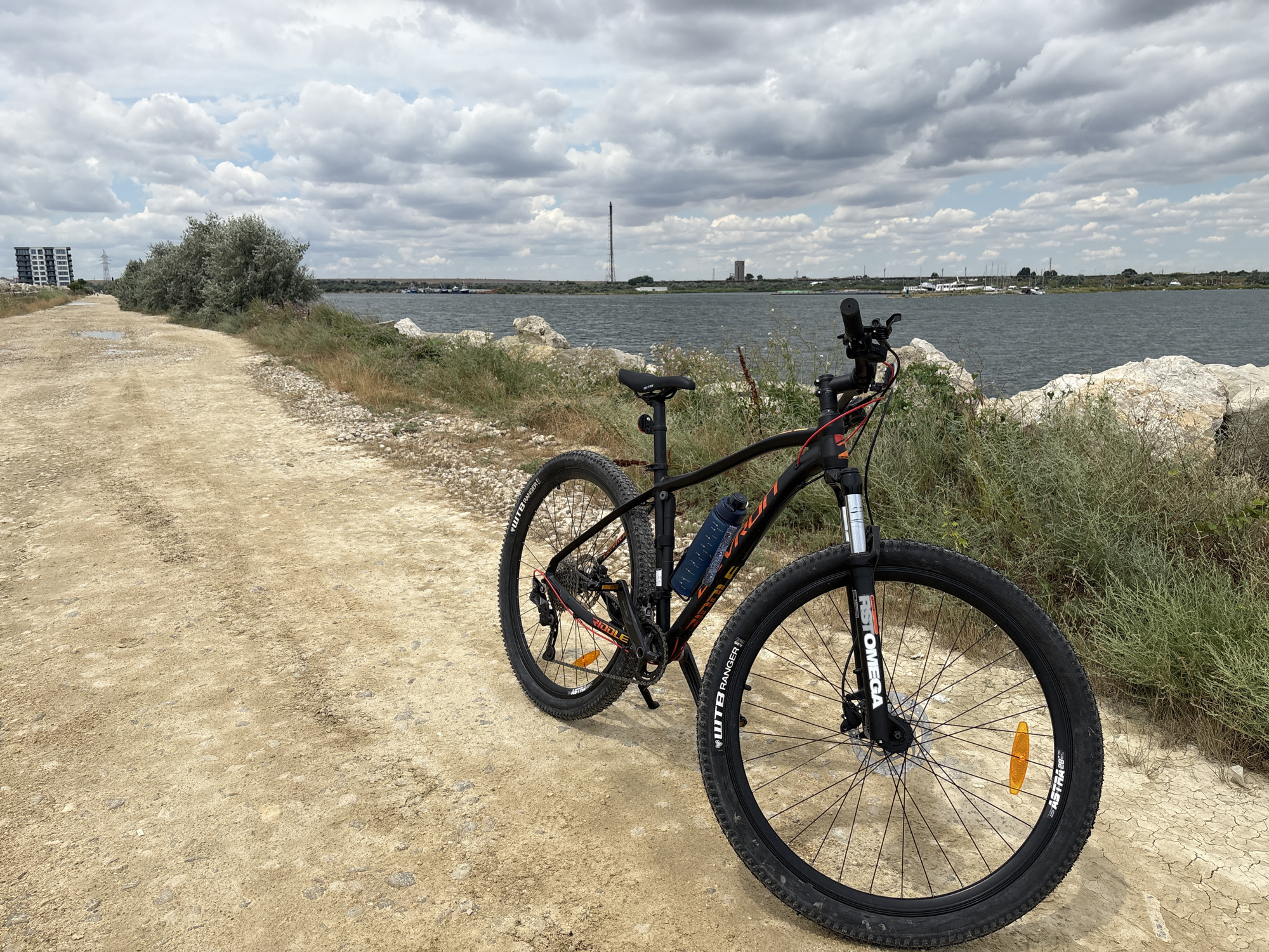 A black mountain bike is parked on a dirt path next to a body of water, likely the Siutghiol Lake, in Năvodari, Romania. The bike has a blue water bottle attached to the frame and orange reflectors on the wheels. In the background, there are clouds in the sky, a few boats on the water, and some buildings on the horizon. The path is made of light brown dirt and gravel, with some patches of dry grass and rocks on the sides. A modern building with multiple floors is visible on the left side of the image, along with a tall transmission tower further in the distance. The overall scene suggests a recreational setting with a focus on outdoor activity.