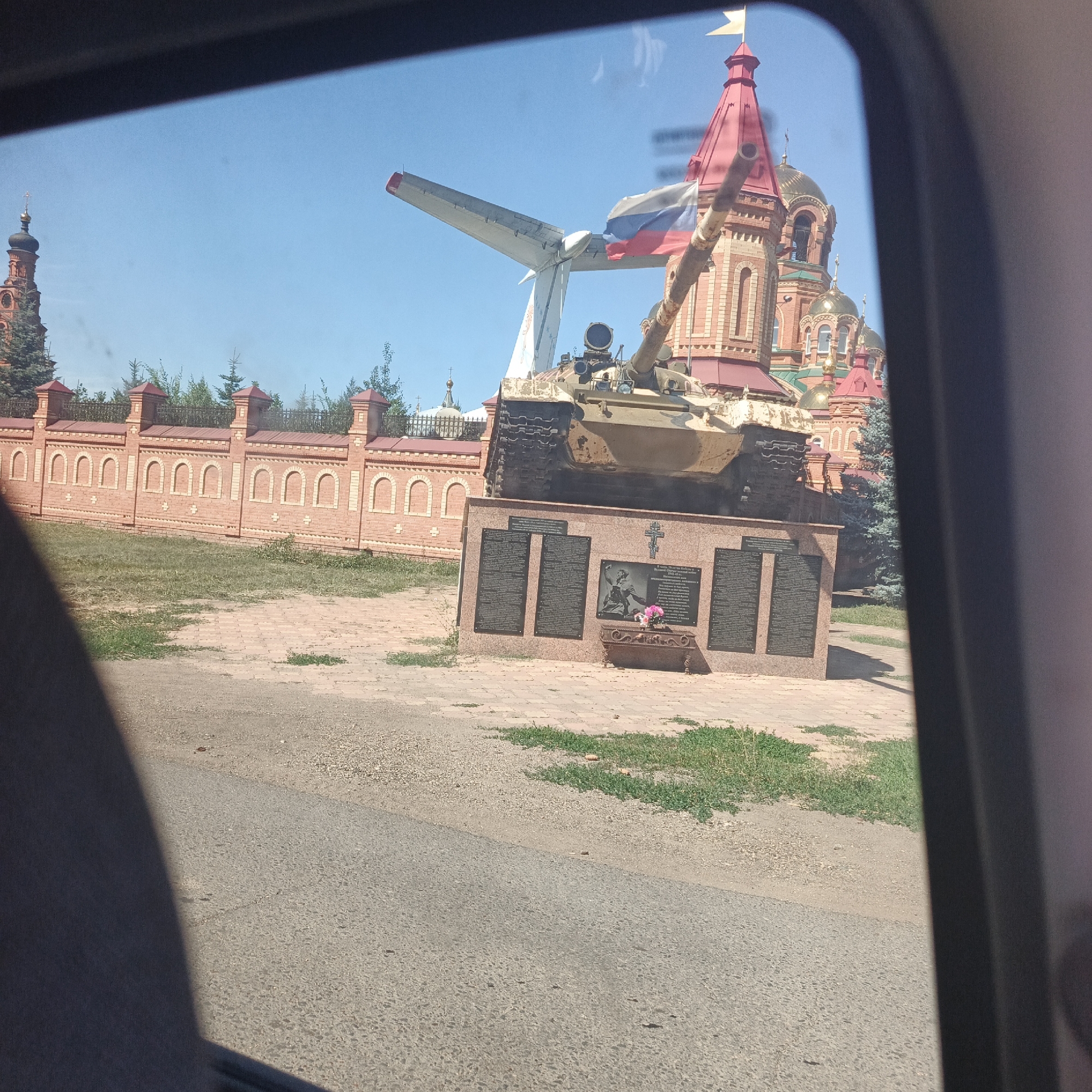 The image, taken from inside a vehicle, shows a war memorial in Saraktash, Russia, featuring a Soviet-era tank and a section of an airplane wing. The memorial is located in front of a brick building with red and gold domes, likely a church. A Russian flag is attached to the airplane wing. The memorial base has several plaques with inscriptions, and a small arrangement of pink flowers sits on a ledge. A brick wall with arched windows runs along the perimeter of the site, behind which trees are visible. The ground is paved with brick and gravel, and a paved road is in the foreground. The sky is clear and blue.