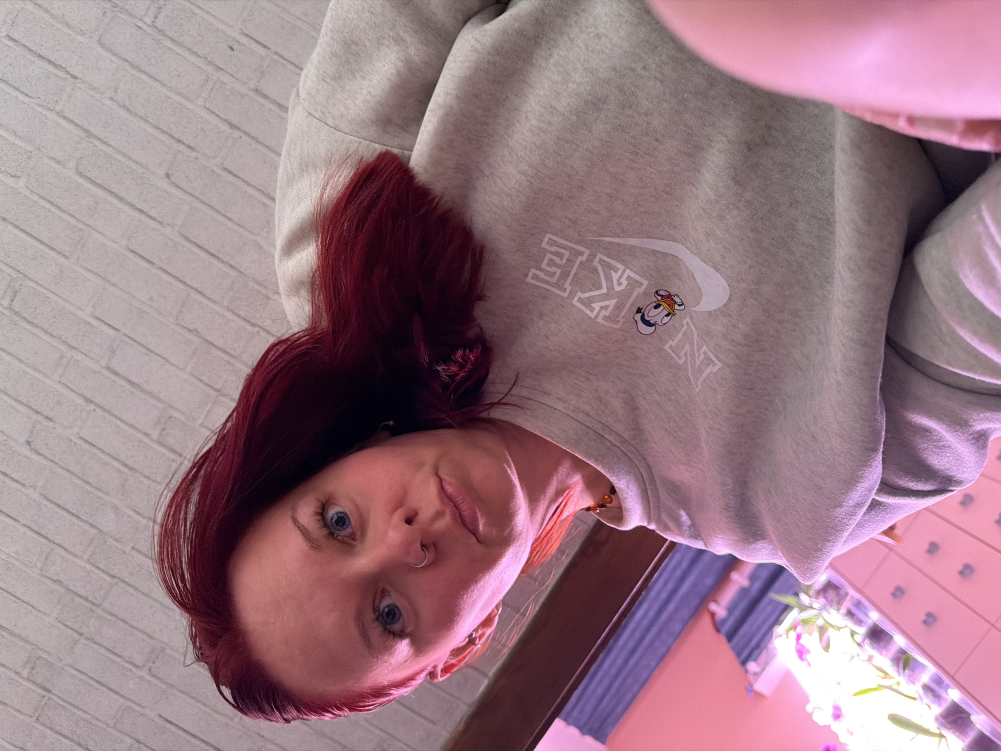 A fair-skinned female with blue eyes and long, vibrant red hair looks directly upward into the camera. She wears a light grey crewneck sweatshirt featuring a white "NIKE" logo with a small cartoon character (Donald Duck) next to the 'N'. A silver hoop nose ring is present on her right nostril, and small silver hoop earrings are visible on both ears. An amber-colored beaded necklace is worn around her neck. Her head is tilted, and her lips are slightly pursed. The upper background consists of a light-colored, brick-patterned wall. To the lower left, an indoor plant cultivation setup is visible, featuring multiple small plants on a white surface, illuminated brightly by a pink-purple light source. A dark horizontal structure is present above the plants. A section of a light pink, fabric-covered surface, consistent with a leg, is visible in the lower right foreground. The scene is illuminated by artificial pink-purple light. This image is observed in Chaikovsky, Russia.